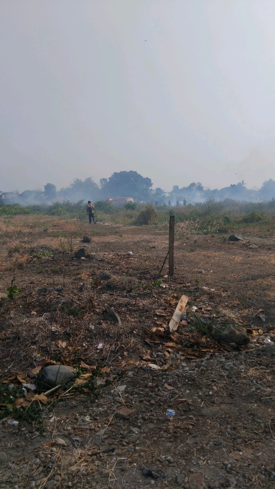 The image depicts an outdoor scene in Tegal, Indonesia, characterized by a hazy, smoke-filled atmosphere suggesting a fire or burning activity in the distance. The foreground consists of dry, barren land with scattered rocks, debris, and dry vegetation. A single wooden post stands vertically in the middle ground. In the background, obscured by smoke, are silhouetted structures, possibly houses or buildings, and several figures of people. One person is visible in the mid-ground, walking away from the camera, appearing to be dressed in dark clothing. The overall environment appears to be an open field with some vegetation, and the presence of smoke indicates a potential fire or controlled burning operation in the vicinity. The sky is a pale, overcast grey, further contributing to the hazy visibility.