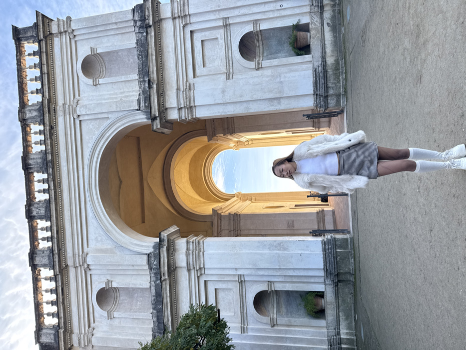 An outdoor, eye-level shot captures a young female standing in the foreground against the imposing stone architecture of the entrance pavilion at Villa d'Este in Tivoli, Italy. The grand structure features a large central arched passage leading through multiple barrel-vaulted sections, illuminated by bright daylight from beyond. A balustrade crowns the building, visible against a partly cloudy sky. Flanking the main archway are two architectural sections, each containing a recessed niche with a potted green plant at its base. The ground in the foreground is a light-brown, unpaved surface. The female subject is positioned slightly right of center, facing left with her head turned slightly to her right. She wears a white ribbed top, a grey and white plaid mini-skirt, sheer dark hosiery, white knee-high socks, and white sneakers. A cream-colored faux fur coat is draped over her shoulders. A green-leafed tree branch is partially visible on the far left.