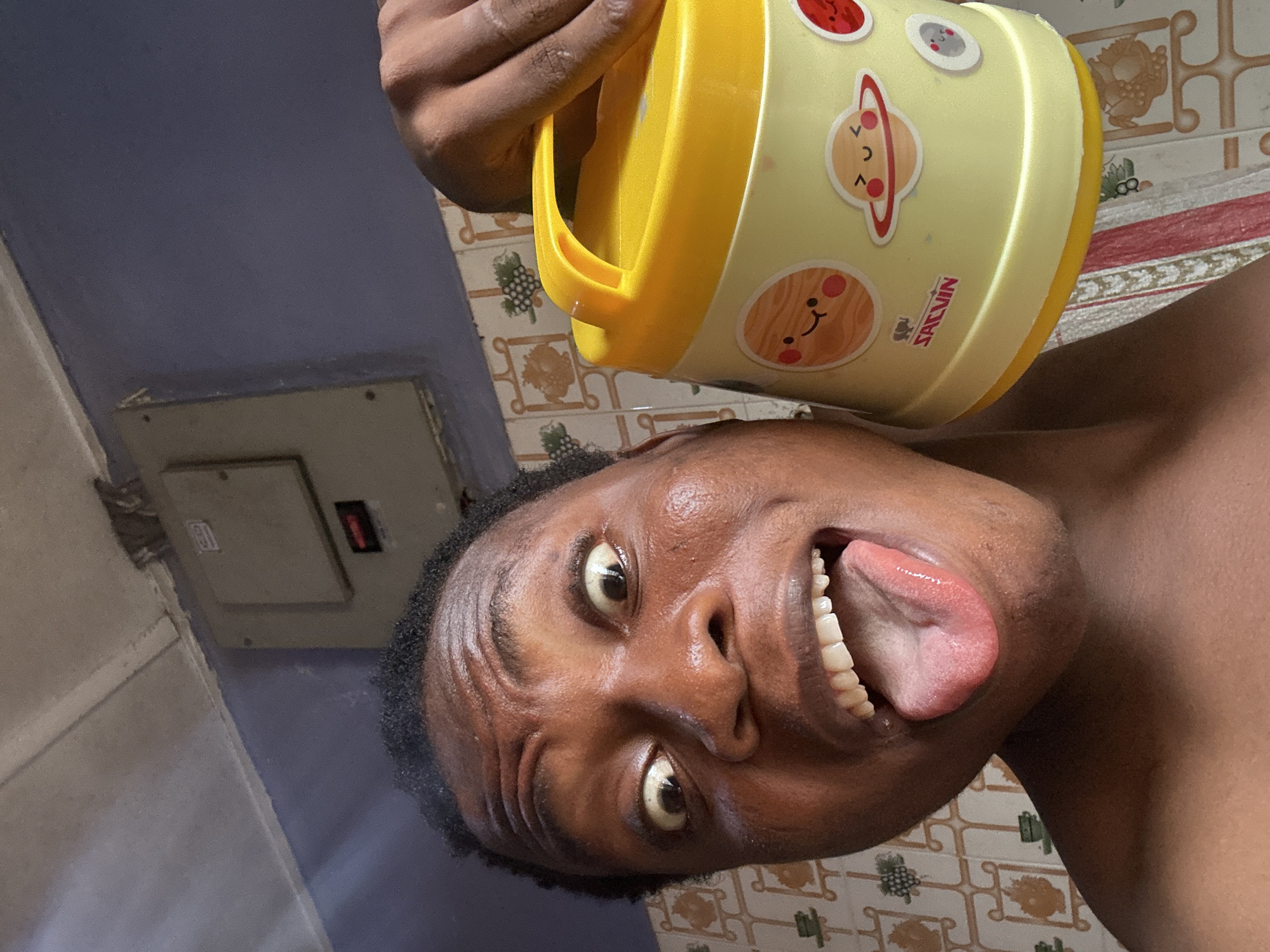 A person with dark skin is holding a yellow plastic container with cartoon planet stickers above their head, with their tongue sticking out. The background consists of patterned bathroom tiles and a blue painted wall with a light-colored electrical panel.