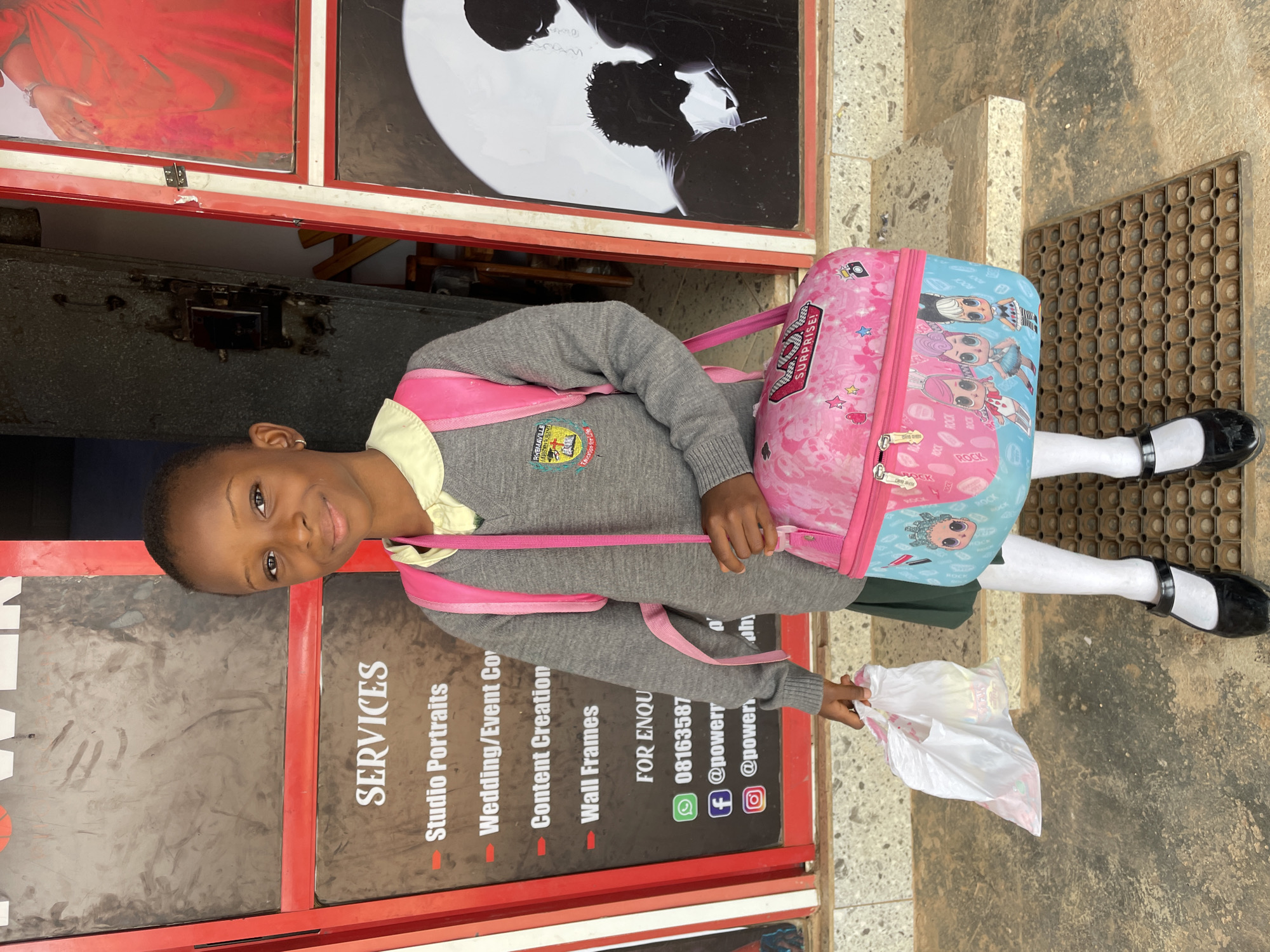 A young Black girl in a school uniform stands in front of a business establishment, holding a plastic bag. She wears a gray sweater with the RhemaVille Academy logo, a green skirt, white tights, and black shoes. A pink and blue backpack is slung over her shoulder.
The girl is smiling and looking at the camera. The business behind her advertises "SERVICES" for studio portraits, wedding/event coverage, content creation, and wall frames. Contact information includes a phone number (08163587), a WhatsApp icon, and Facebook and Instagram handles (@powerr).
The location is determined to be in Barakin Akawo, Nigeria, based on the provided context, particularly the phone number with the 081 dialing code, commonly used in Nigeria.