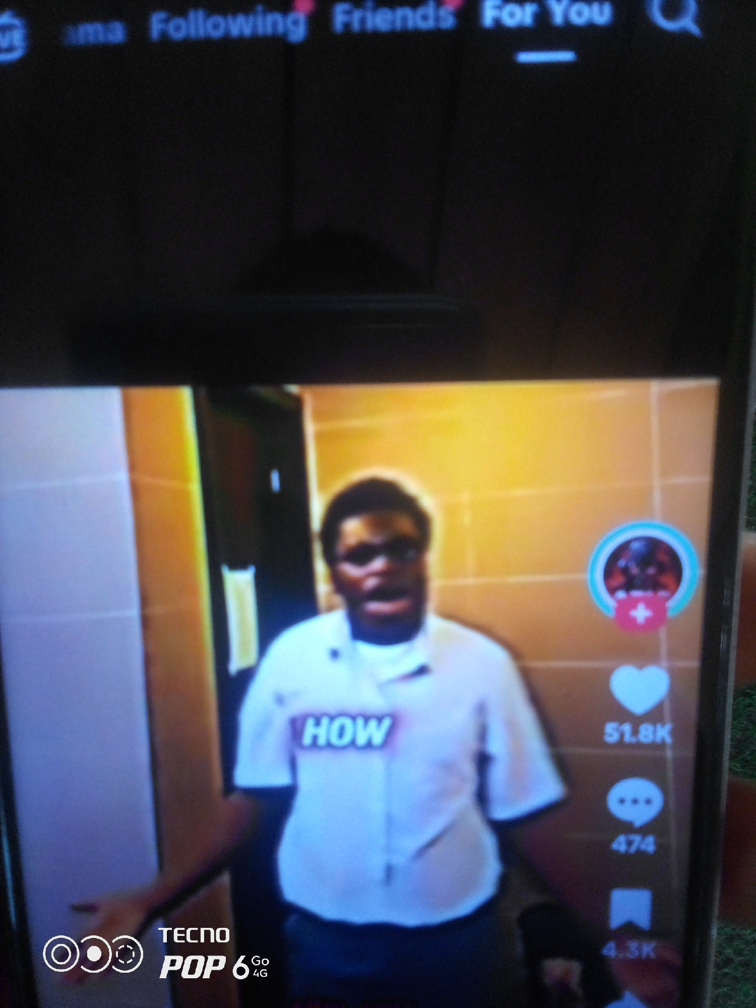 A close-up, slightly blurry photograph captures a person's face and upper body on a smartphone screen. The person, appearing to be of African descent with dark skin and short, curly hair, is looking directly at the camera with an open mouth, suggesting they are speaking or singing. They are wearing a light-colored shirt with "HOW" written across the chest in a contrasting color. The background on the screen is an interior setting with warm, yellowish walls.
On the right side of the phone screen, social media engagement icons are visible: a profile picture with a plus sign, a heart icon with "51.8K" below it, a speech bubble icon with "474" below it, and a bookmark icon with "4.3K" below it. The bottom left corner of the phone screen displays "TECNO POP 6 Go 4G" in white text. The image does not contain any information or visual cues that would allow for its localization to Howayi, Nigeria, or any other specific city.