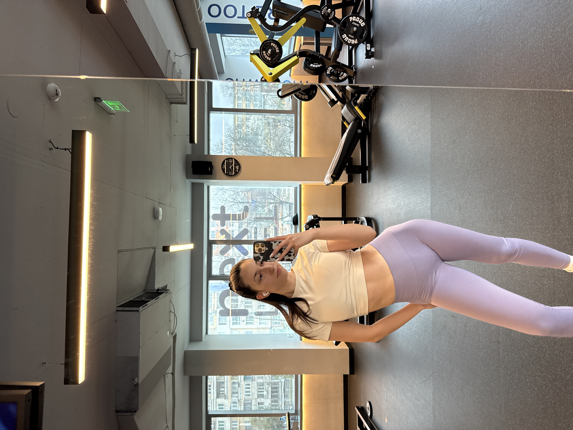The image depicts a young woman taking a selfie in a gym. She is wearing a cream-colored crop top and light purple leggings. The gym has large windows, a clock on the wall, and workout equipment. Based on the window view, the location appears to be in an urban area with buildings and trees. Specific landmarks are not visible to confirm Sofia, Bulgaria. The woman is holding a smartphone and appears to be taking a picture of herself in the mirror. She is centered in the frame, with gym equipment and architecture in the background.