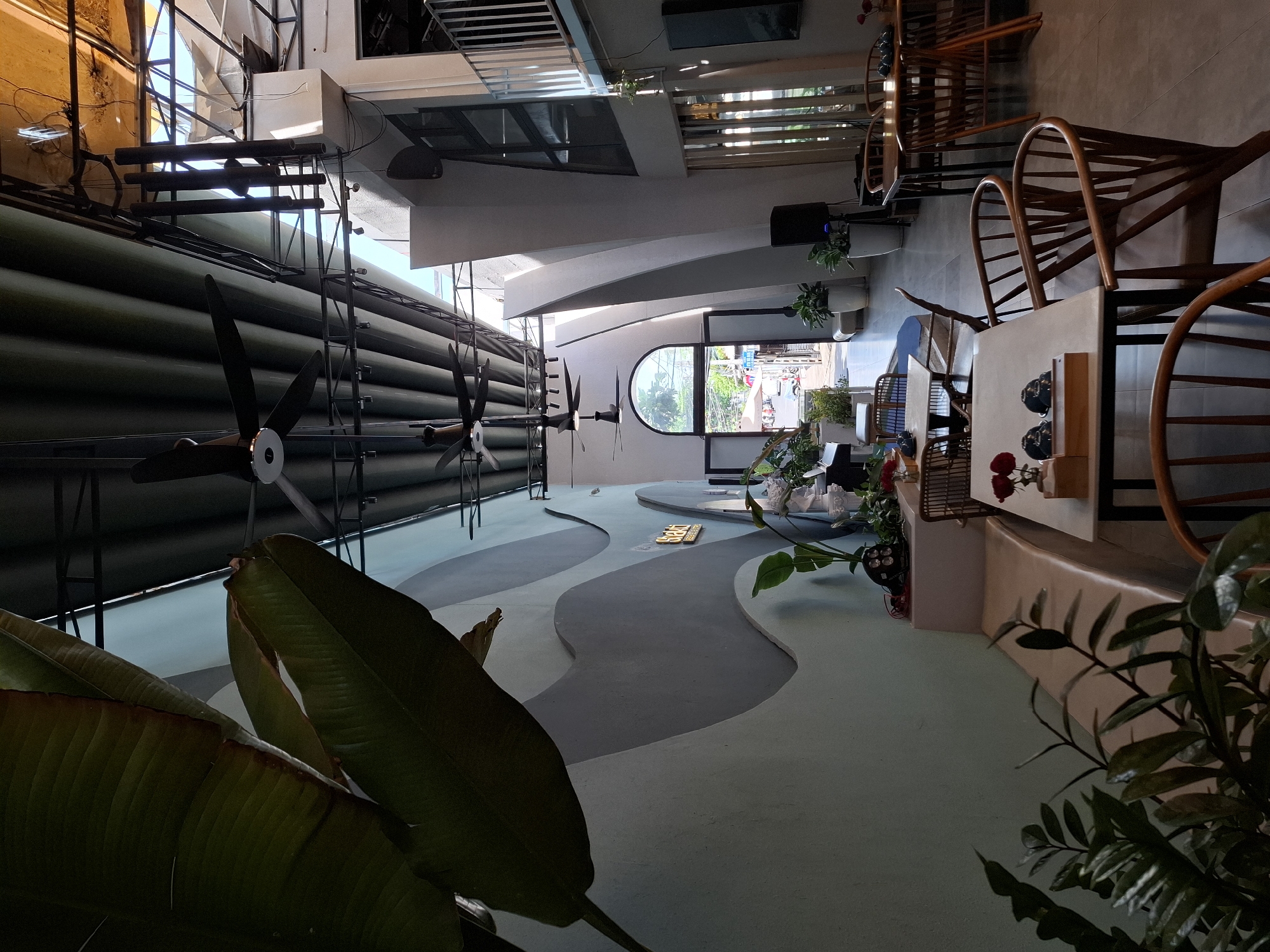 The image depicts the interior of a cafe located in Nha Trang Ward, Vietnam. The cafe features a modern, open-plan design with high ceilings and exposed structural elements. Large tropical plants, including banana leaves, are prominently displayed, adding a touch of nature to the space. The walls are decorated with a wave-like pattern in shades of blue and grey.
Several ceiling fans are visible, suggesting a warm climate. The seating area includes a mix of wooden tables and chairs, as well as a long, curved bench. In the background, a large arched doorway opens to the outside, revealing a street scene with traffic and pedestrians.
The cafe appears to be empty of customers, with only a few scattered decorative items like vases of red roses on the tables. The lighting is a mix of natural sunlight streaming in from the open entrance and artificial lights. The overall atmosphere is bright and airy, with a focus on natural materials and a contemporary aesthetic.
