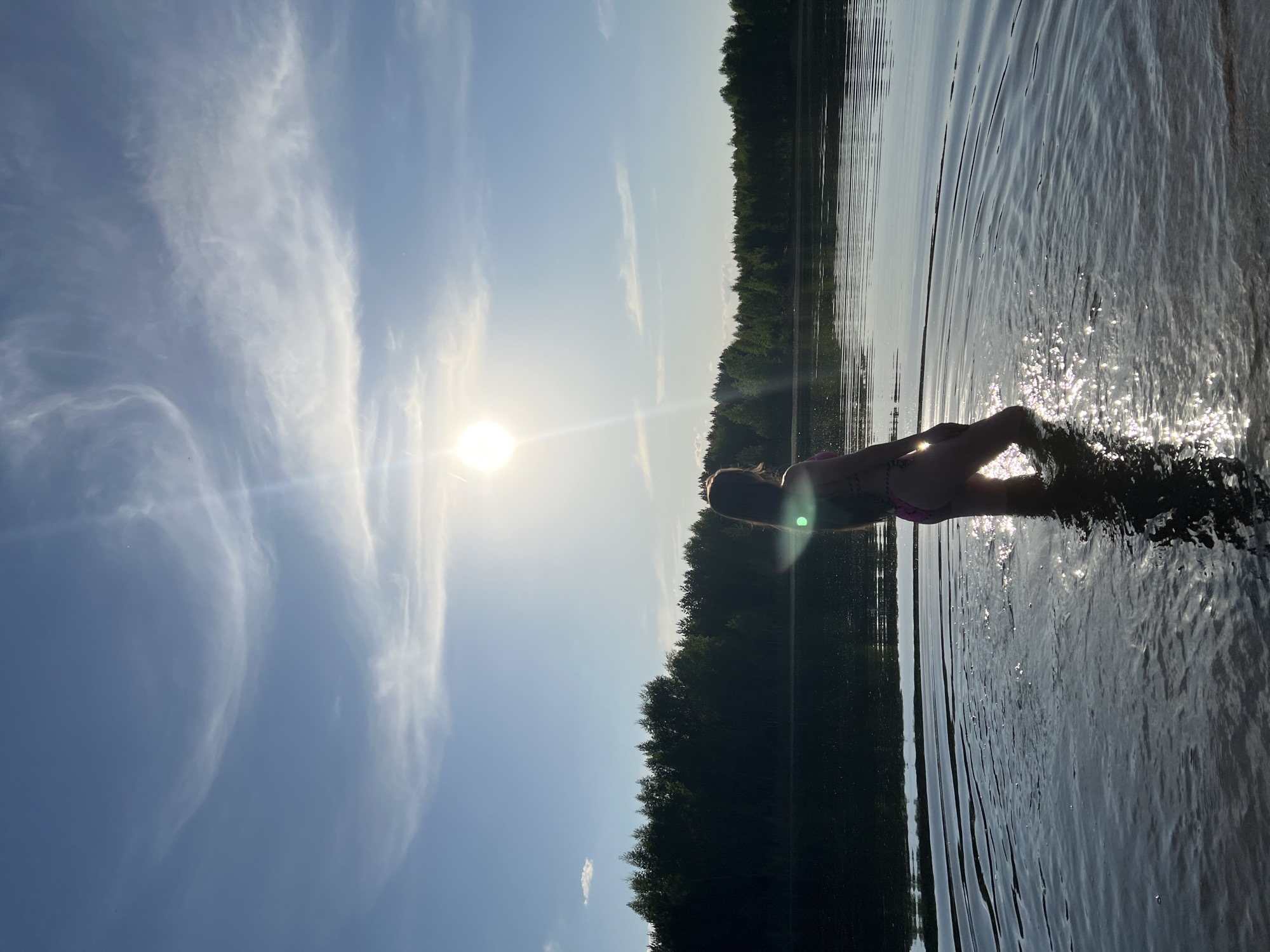 A horizontal shot shows a person in the water. The upper body is partially out of the water. The person's skin is dark, but is visible enough to observe a tattoo on the arm. The person is wearing a two-piece bathing suit. The sun is in the upper-left of the shot, reflecting in the water. In the background is a dark forest. The sky is pale blue with some whispy white clouds. The location may be in or around Novyi, Russia.