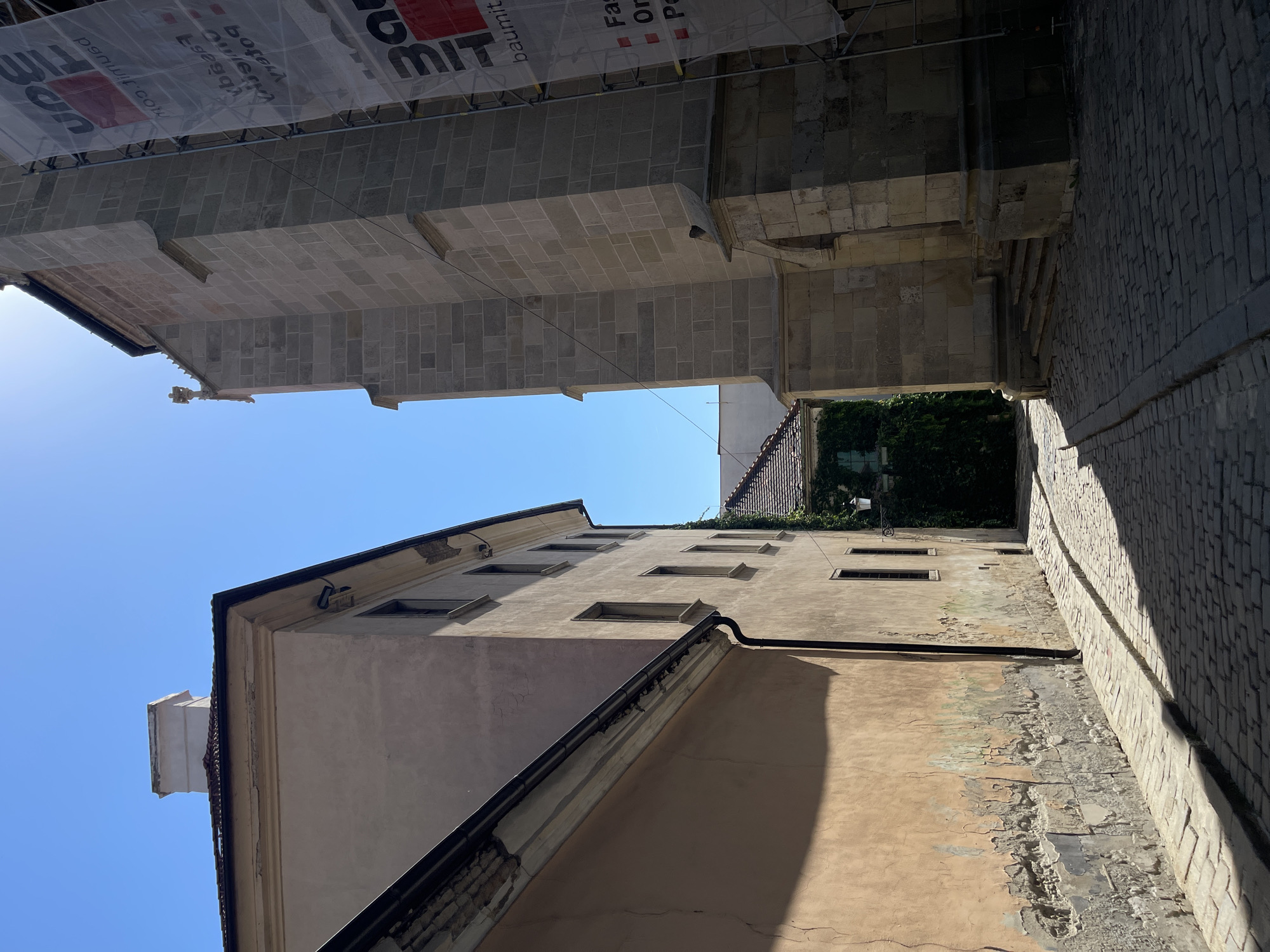 The image depicts a narrow cobblestone street in Bratislava, Slovakia, with historic buildings on either side. On the left is a light pink building with a damaged facade showing peeling paint and a black gutter. The building to the right is a stone structure, possibly a church or cathedral, with scaffolding and a large banner erected on its side. The banner has text and logos, but it is partially obscured. Sunlight casts distinct shadows across the street, indicating a bright, clear day. The sky is a vibrant blue. In the distance, greenery can be seen clinging to a building, adding a touch of nature to the urban scene. The overall atmosphere is one of historical architecture and a quiet, sunny day in the city.