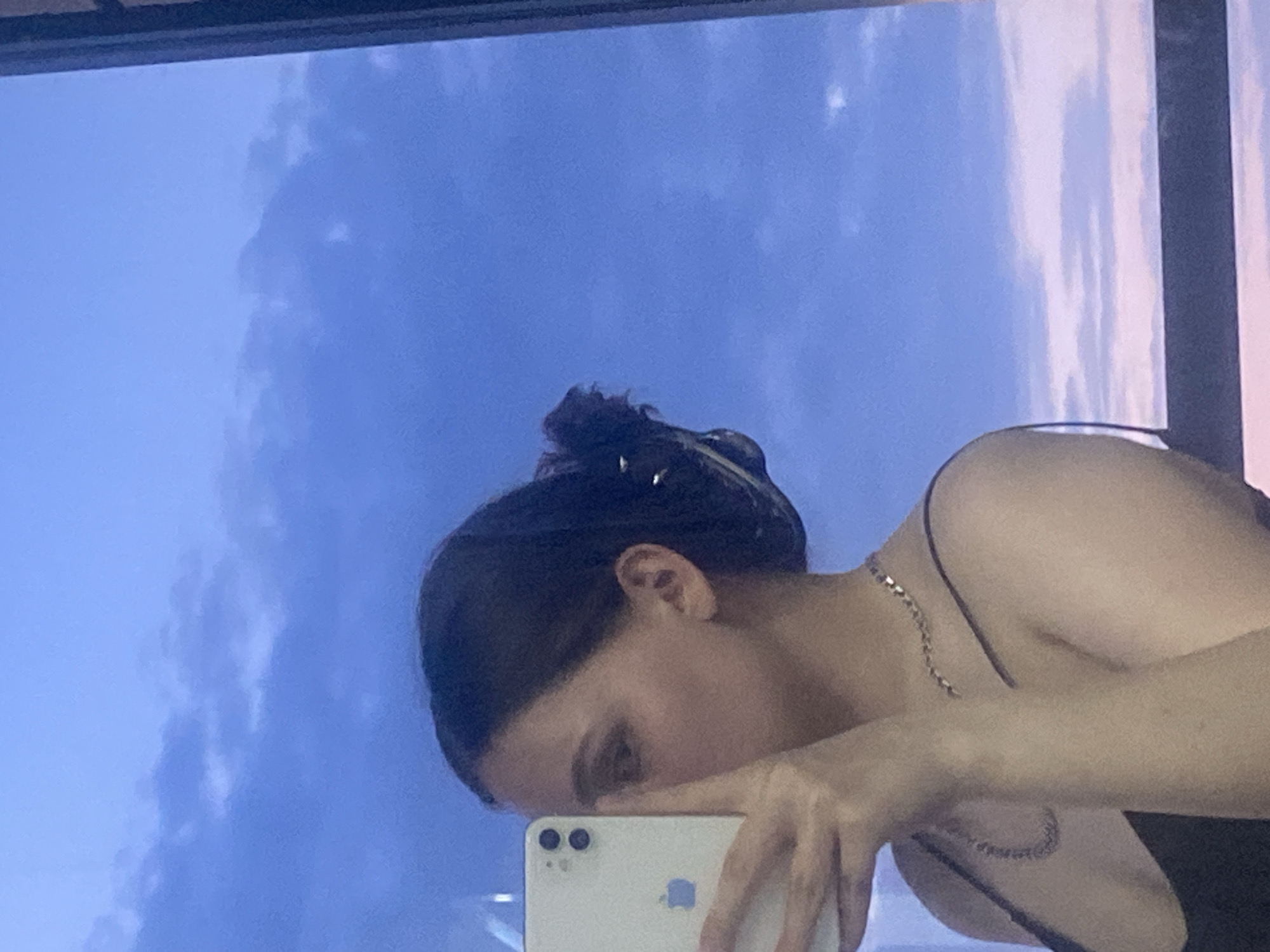 A woman is taking a selfie in front of a window. The window is reflecting a blue sky with clouds. The woman has dark hair pulled back in a bun and is wearing a thin-strapped top. She is holding a white iPhone in her right hand. The image does not contain sufficient information to determine the location as Izhevsk, Russia.