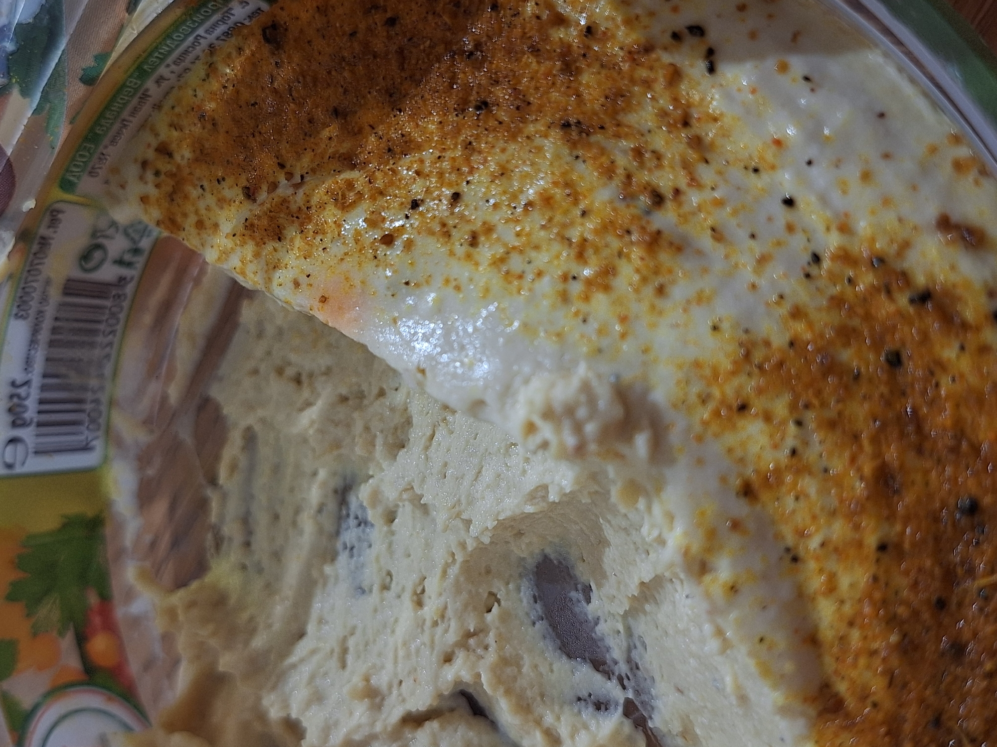A close-up, overhead shot captures a container of creamy food, likely a dip or spread, with a portion of its clear plastic lid peeled back. The main food substance is a thick, light beige or cream color, exhibiting a lumpy texture with darker, irregular pieces embedded within it, possibly indicating ingredients like olives or mushrooms. A smoother, lighter layer covers the surface, which is generously dusted with orange-brown spices, resembling paprika or chili powder, and fine black specks, likely black pepper.
The left side of the image reveals part of the plastic container and its label. The label features a barcode and partially visible text. Notable text fragments include "3200" and "100", along with characters from a non-Latin alphabet, appearing to be Cyrillic or similar. Reversed text "ДОНАС ПОЧТИ" (Donas Pochti, possibly meaning "almost Donas") is visible, suggesting a brand or product name. A price "3200 €" is also faintly discernible. The label's design includes a section with green, white, and a leaf graphic. The lighting is consistent, accentuating the food's texture and the details on the packaging. The scene is static, focusing entirely on the food product.