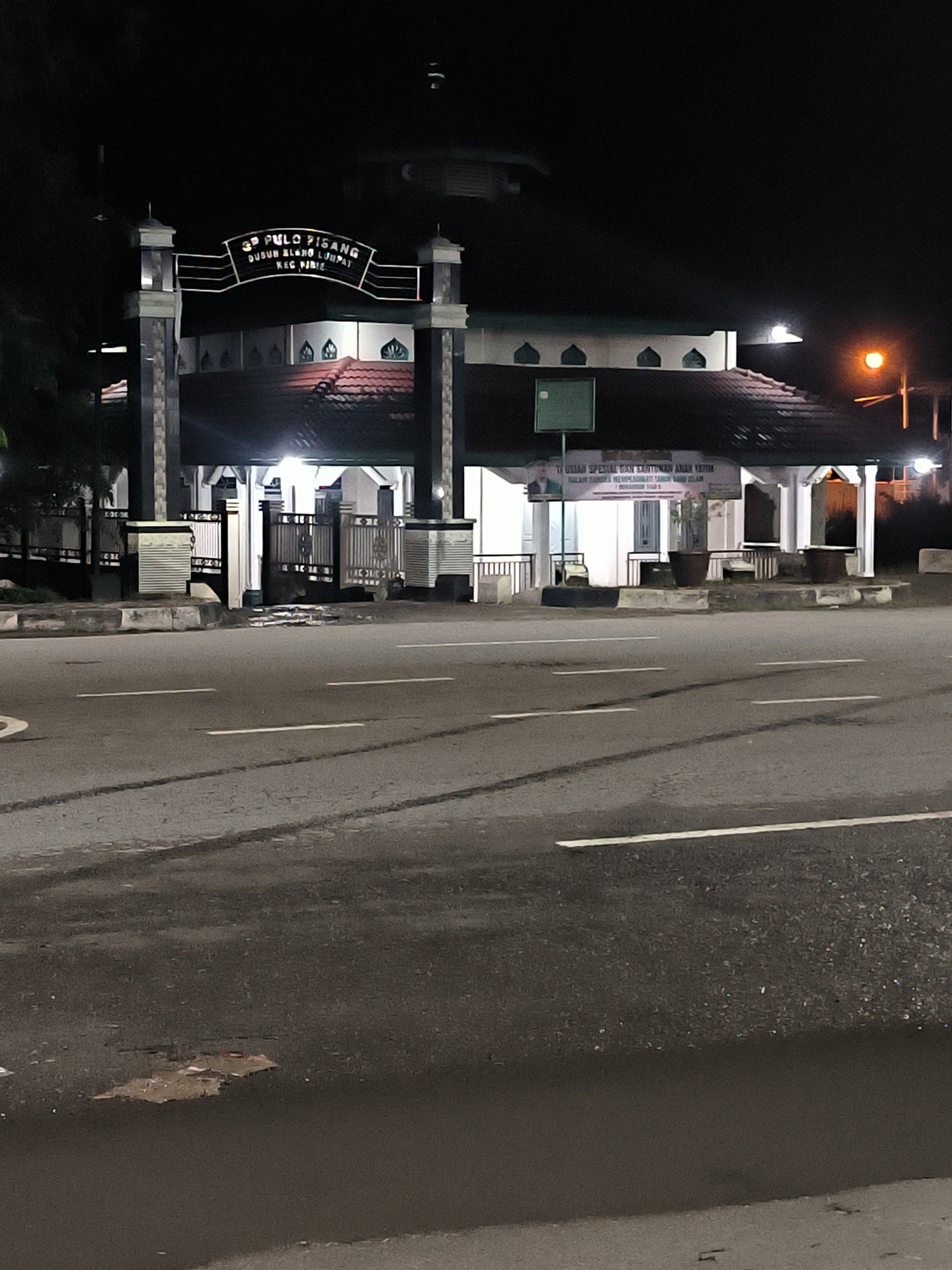 The image shows the entrance to a building at night, likely a mosque or religious center, in Sigli, Indonesia. A prominent sign above the entrance reads "39 PULO PISANG DUSUN BLANG LUMPAT KEC. JURIE." This indicates the location as Dusun Blang Lumpat, Pulo Pisang, in the Jurie sub-district of Sigli. The building has a distinctive architectural style with a red tiled roof and arched window-like features. White gates and pillars mark the entrance, with some decorative elements visible. A street with white lane markings runs in front of the building. A lamppost with an orange light is visible to the right of the entrance. The scene is illuminated by artificial lights, creating shadows and highlighting the building's features against the dark night sky. There are no people visible in the frame.