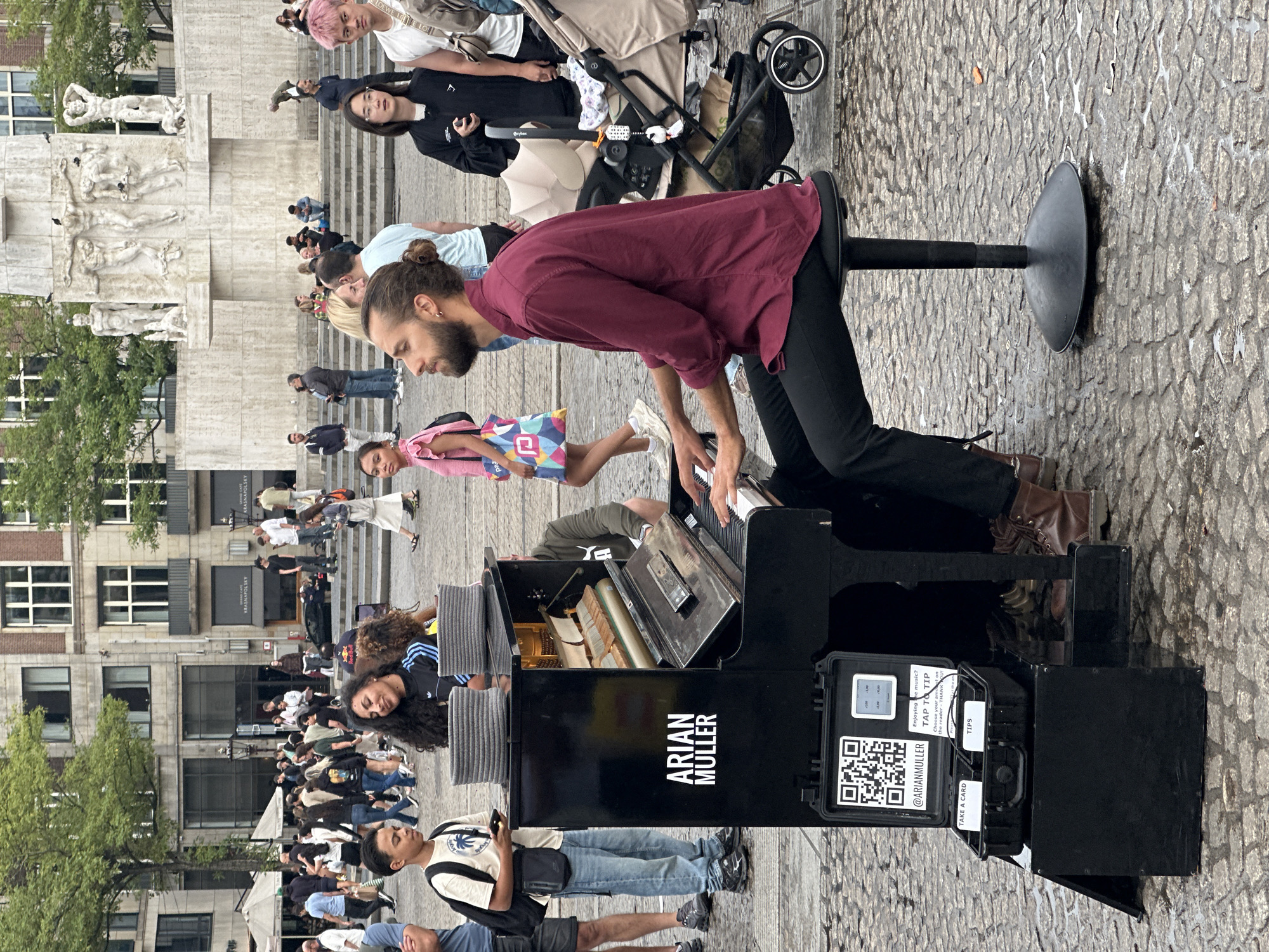 The image shows a street scene in Dam Square, Amsterdam, Netherlands. In the foreground, a man with a beard and his hair tied in a bun is playing a black portable piano. He is wearing a maroon shirt and black pants. Next to the piano is a stool he is sitting on. In front of the piano, there is a black case with a QR code, a payment terminal, and two compartments labeled "TAKE A CARD" and "TIPS". The piano itself has "ARIAN MULLER" written on its side. Behind the pianist, a young boy in a t-shirt and jeans is looking at his phone. Further back, a crowd of people are walking around, and a monument with sculptures is visible in the background. The ground is paved with cobblestones.
