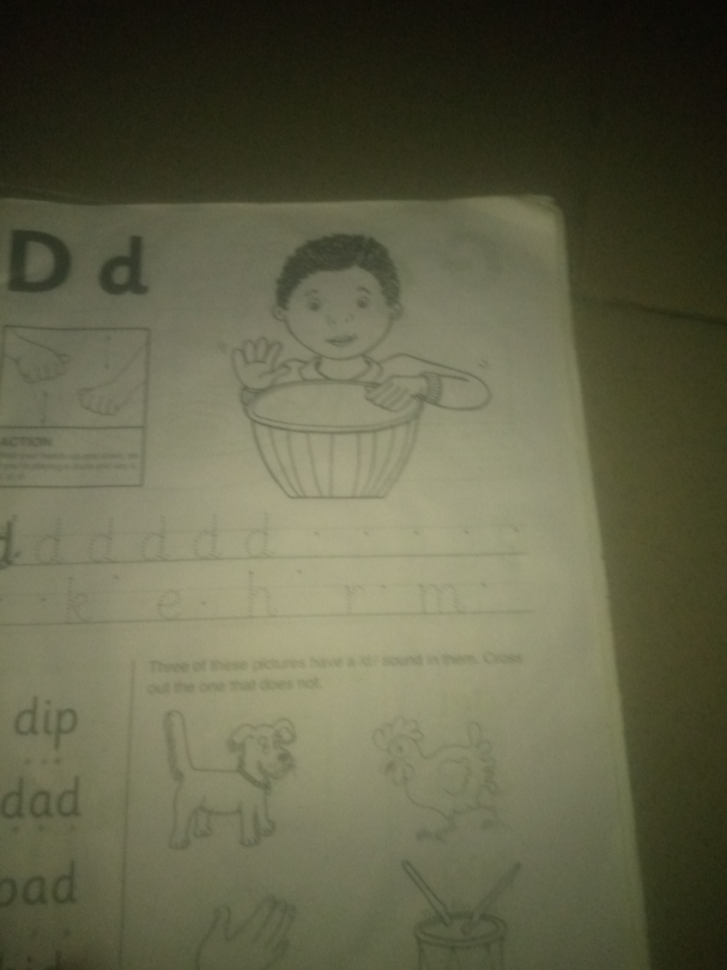 The image shows a page from a children's educational worksheet. At the top left, there are large block letters "D d". To the right of the letters, there is a line drawing of a child, likely a boy, smiling and holding a bowl with both hands. Below the letters "D d", there are lines for practicing writing lowercase letters. The first line shows a tracing of the letter "d" repeated multiple times. The second line shows the letters "k", "e", "h", and "r" traced.
On the bottom left of the page, there are words written in a larger font: "dip", "dad", and "bad".
On the right side of the page, there are three line drawings of objects. The top drawing is of a dog. The middle drawing is of a chicken. The bottom drawing is of a drum with drumsticks. Above these drawings, there is text that reads: "Three of these pictures have a /d/ sound in them. Cross out the one that does not."
The image appears to be a photograph taken in poor lighting conditions, with a dark, out-of-focus background. There are no discernible elements that would place the image in Jauro Yinu, Nigeria. The content is purely educational material.