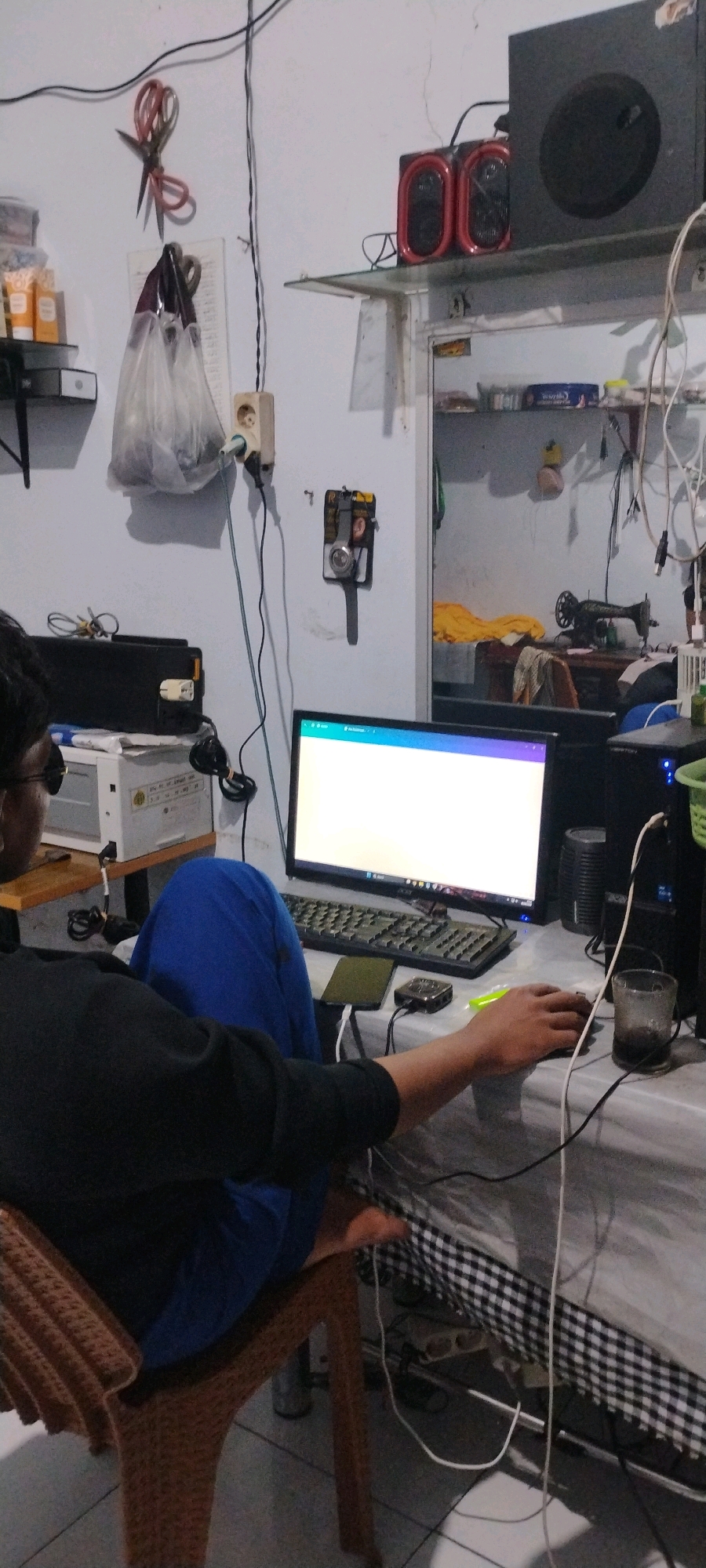 A person is seated in a chair, facing a computer monitor. The person is wearing a black long-sleeved shirt and blue pants. They are using a mouse and keyboard. The computer setup is on a table with a checkered cloth draped over it. In the background, there are shelves with various items, including speakers, a sewing machine, and what appear to be toiletries. A power strip with multiple outlets is visible on the wall, with various cords plugged into it. A pair of scissors is hanging on the wall, attached to a hook. A plastic bag filled with items is also hanging near the scissors. The scene appears to be indoors, in a residential or small workshop setting. The overall impression is one of a cluttered workspace.