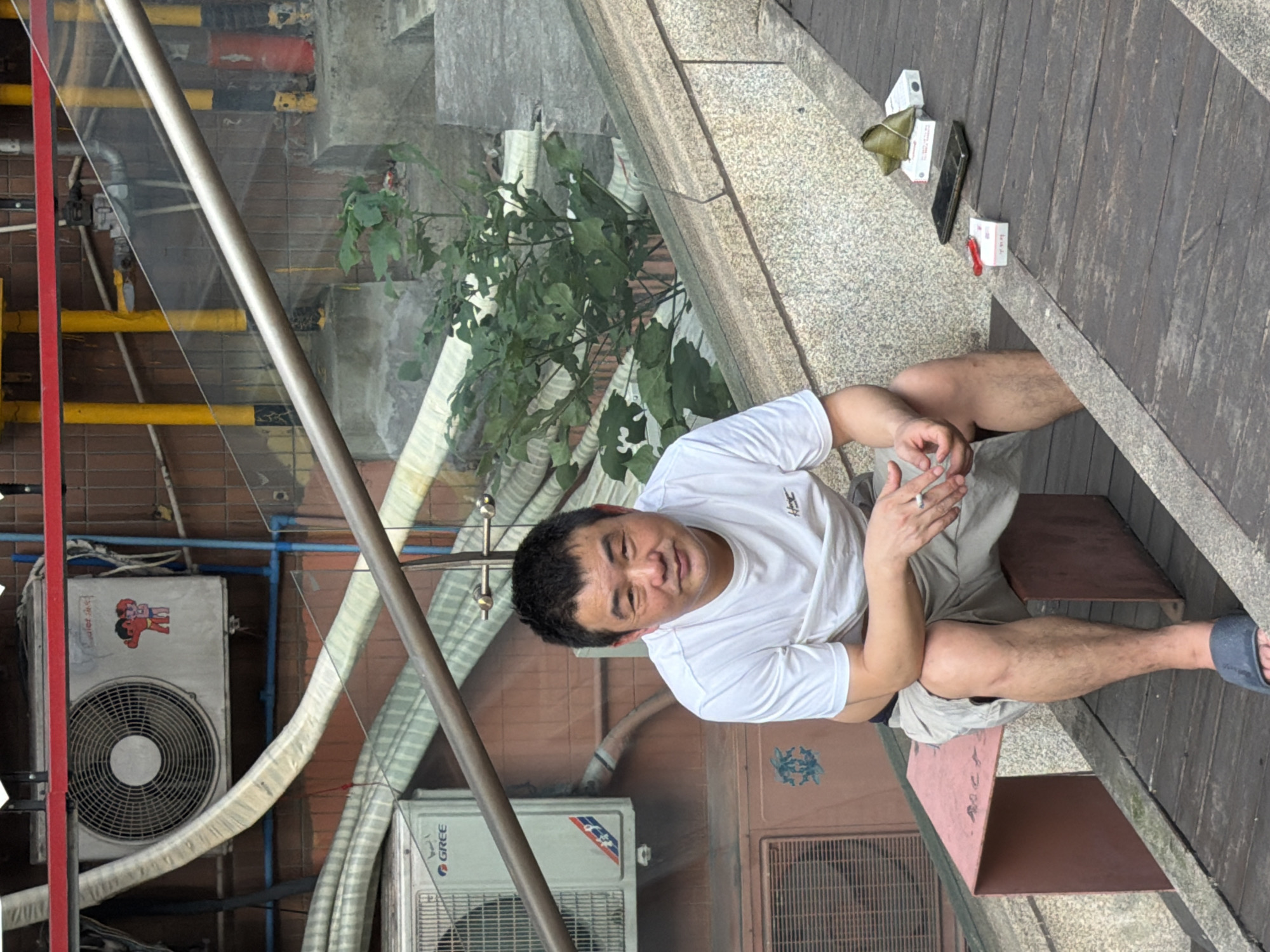 A middle-aged East Asian man sits on a pink bench on an outdoor staircase in Yuzhong District, China. He is wearing a white t-shirt and light-colored shorts, with dark blue sandals on his feet. He holds a lit cigarette in his right hand and appears to be looking towards the camera. A pack of cigarettes and a cell phone are on the wooden steps to his right. Behind him, there are air conditioning units, pipes, and a glass railing. The environment appears to be urban, with a mixture of concrete, brick, and wood elements.