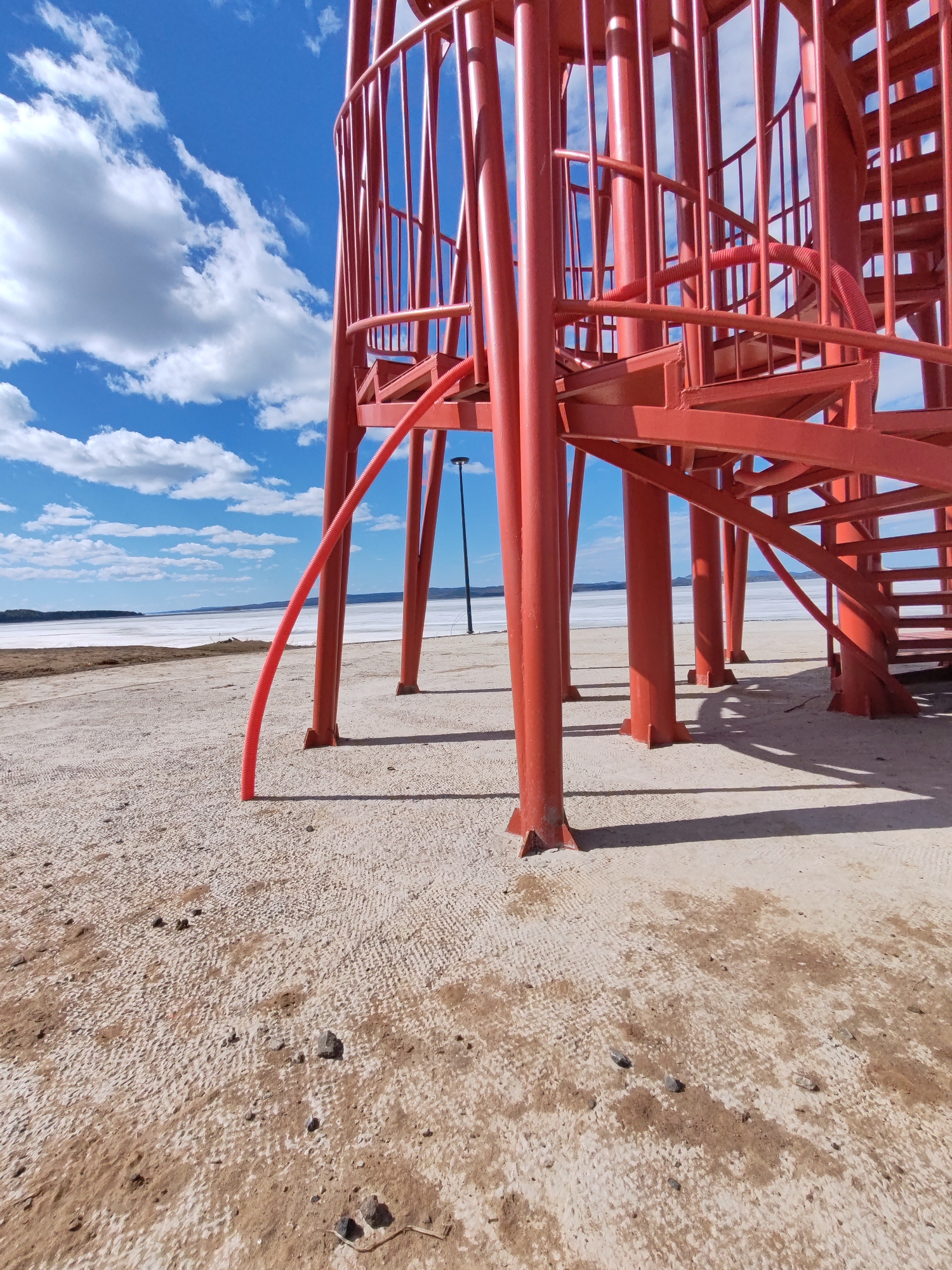 The image depicts a red metal structure with spiral stairs, possibly an observation tower, situated on a sandy or gravelly ground. A red corrugated pipe runs down from the structure to the ground. In the background, a lake or body of water extends to the horizon, with a blue sky and scattered clouds above. A lamppost is visible in the background between the structure's supports. The location is Ust-Ilimsk, Russia.