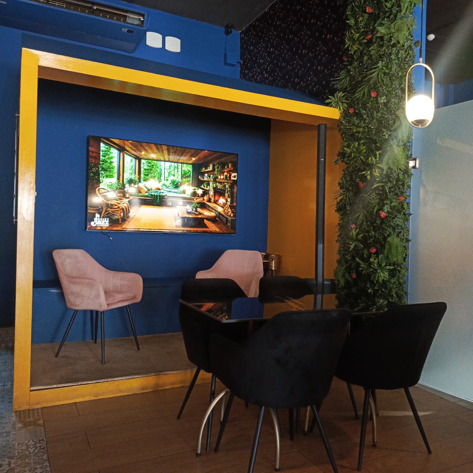 This image depicts an interior scene, likely a cafe or restaurant, characterized by a vibrant blue wall as the primary backdrop. A large television screen mounted on this wall displays an image of a cozy living room with a fireplace and large windows overlooking a forest. In front of the television, a plush pink armchair with thin metal legs is placed on a raised, beige platform, enclosed by a bright yellow frame. To the right of the pink chair, a black, plush armchair is visible, partially obscured by a modern glass-topped table with black chairs surrounding it. A tall, artificial green plant installation, adorned with red artificial flowers, stands to the right of the table, adding a touch of nature to the space. A contemporary pendant light hangs above the plant. The floor is a mix of wooden planks and patterned tiles. The overall ambiance suggests a modern and inviting space. This image was taken in the city of Caracas, Venezuela.