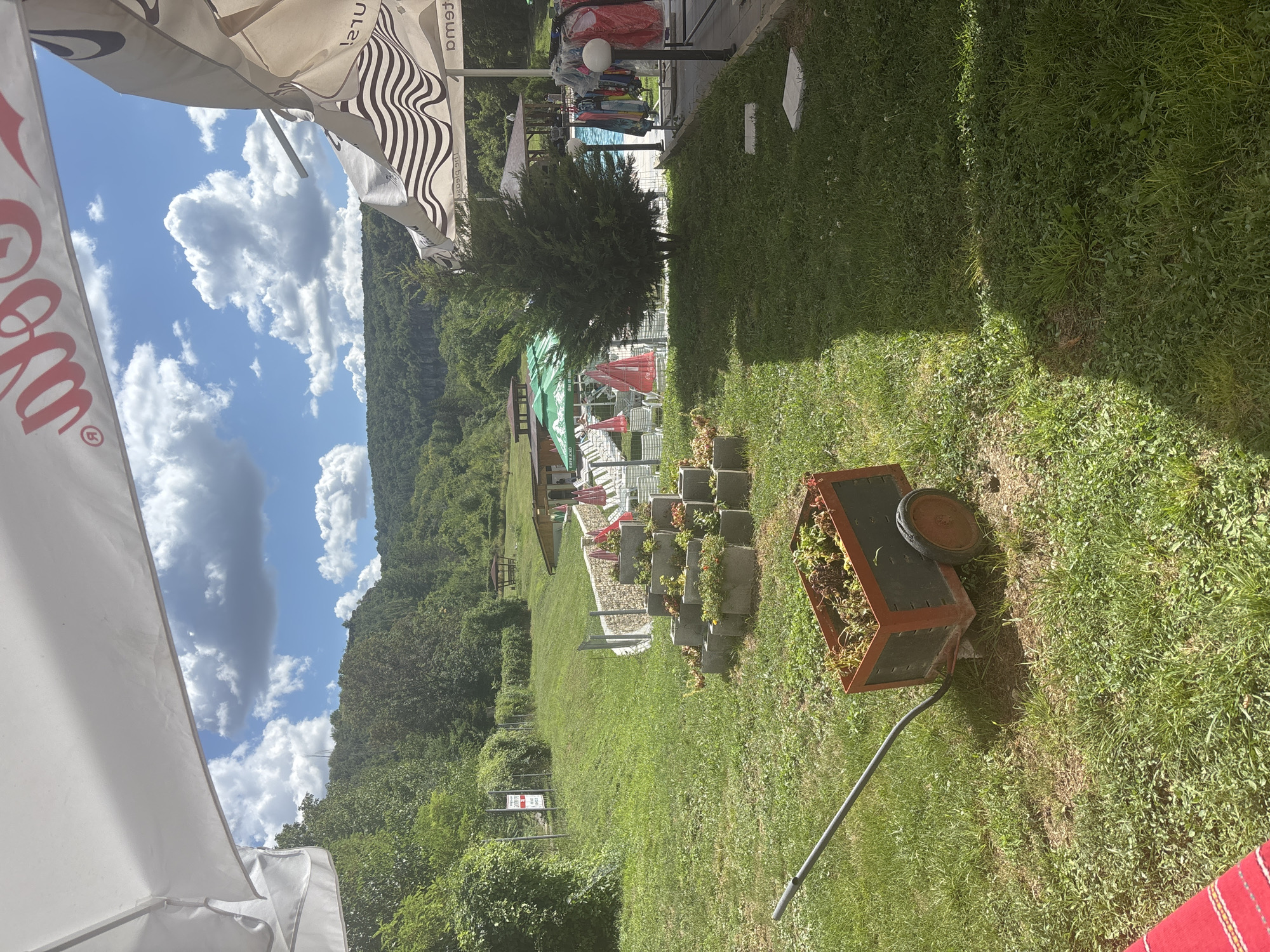 The image depicts an outdoor scene, likely a resort or a recreational area, with elements suggesting it is located in Krushuna, Bulgaria. In the foreground, a red-wheeled cart filled with foliage is positioned on a grassy lawn. Behind the cart, a tiered planter made of concrete blocks holds various plants. Beyond this, a swimming pool is visible, with lounge chairs and umbrellas surrounding it. In the middle ground, there are more lounge chairs and umbrellas, along with a covered gazebo. The background is dominated by a lush, tree-covered hillside under a bright blue sky with scattered white clouds. The presence of a sign with Bulgarian text further points to the location being in Bulgaria. The overall impression is one of a well-maintained outdoor space, possibly a tourist attraction, given the amenities and the scenic natural surroundings.