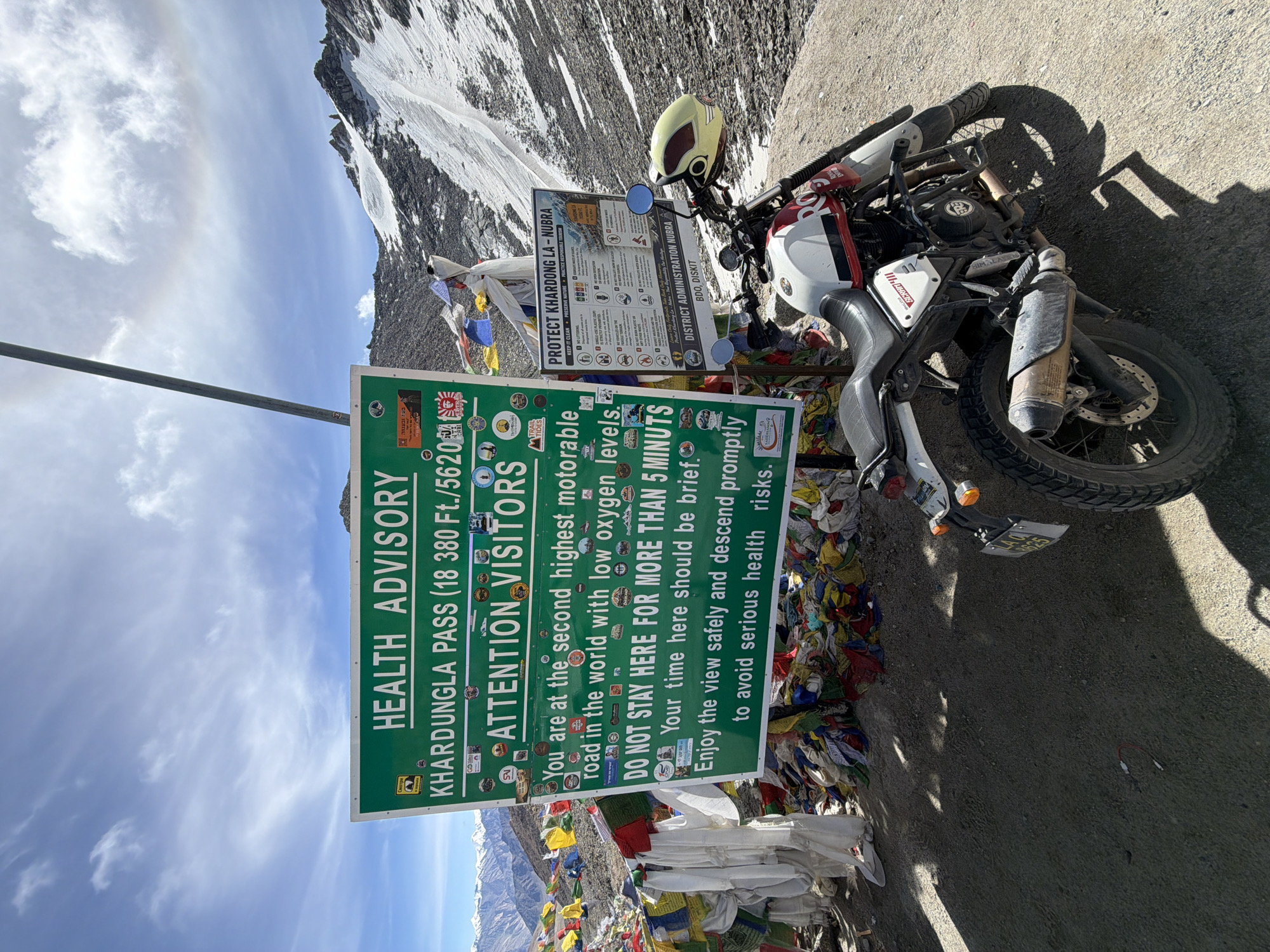The image shows a mountain pass with prayer flags and a large green sign that reads "HEALTH ADVISORY KHARDUNGLA PASS (18 380 Ft./5620m) ATTENTION VISITORS". The sign provides advice on low oxygen levels and recommends limiting time spent at the location to avoid health risks. To the right of the main sign, there is another sign titled "PROTECT KHARDONG LA-NU BRA". A white, red, and black Royal Enfield motorcycle is parked in front of the signs. The background features snow-capped mountains and a cloudy sky with a visible rainbow. The ground is a mix of dirt and gravel. The scene appears to be at a high-altitude location.