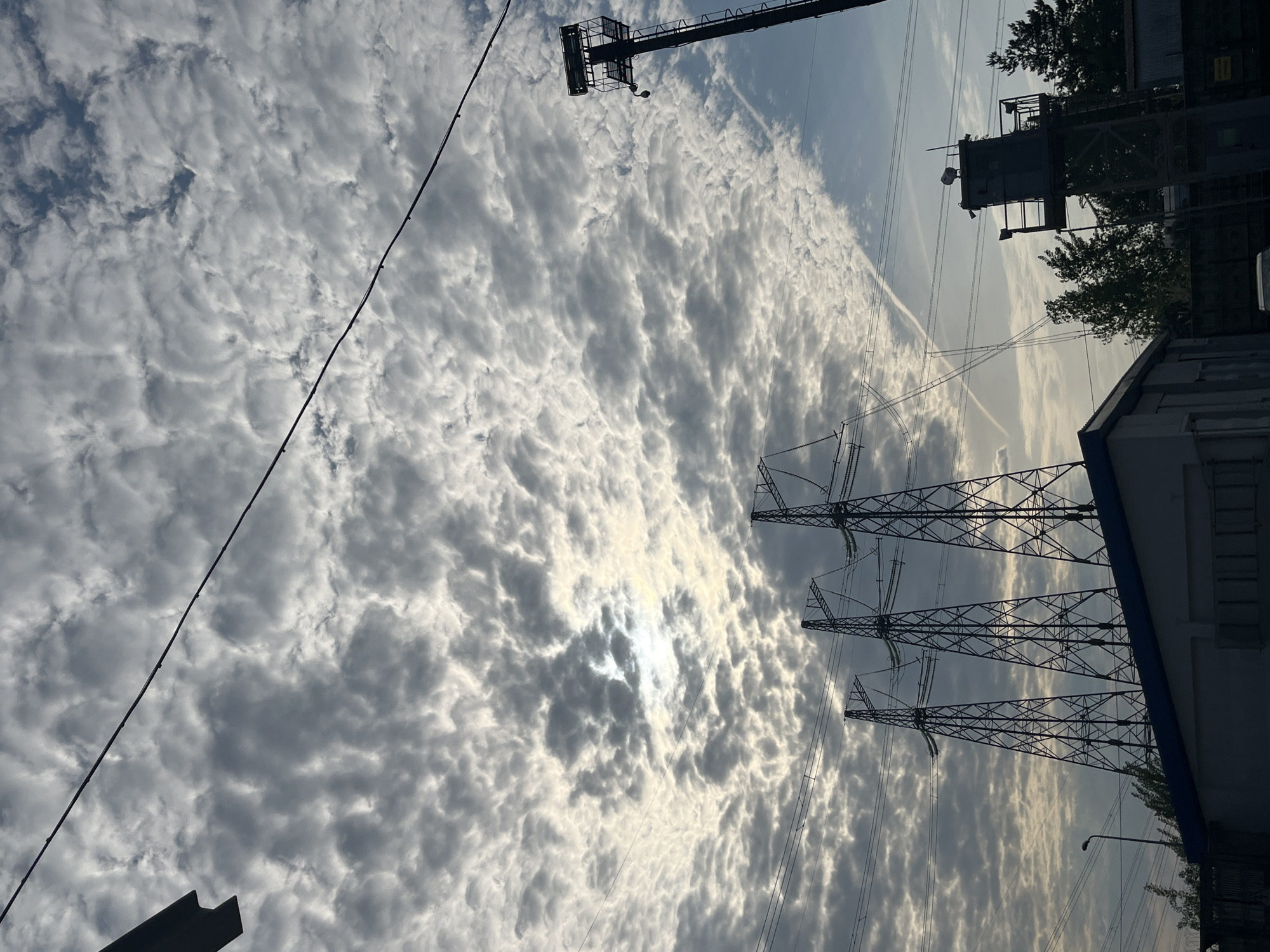 The image shows a view of the sky with altocumulus clouds over Chaikovsky, Russia. The sun is partially obscured by the clouds. Three high-voltage electricity pylons with power lines stretch across the frame. Part of an industrial building is visible on the right side. Other power transmission towers and equipment can be seen in the background. A single contrail cuts through the sky.