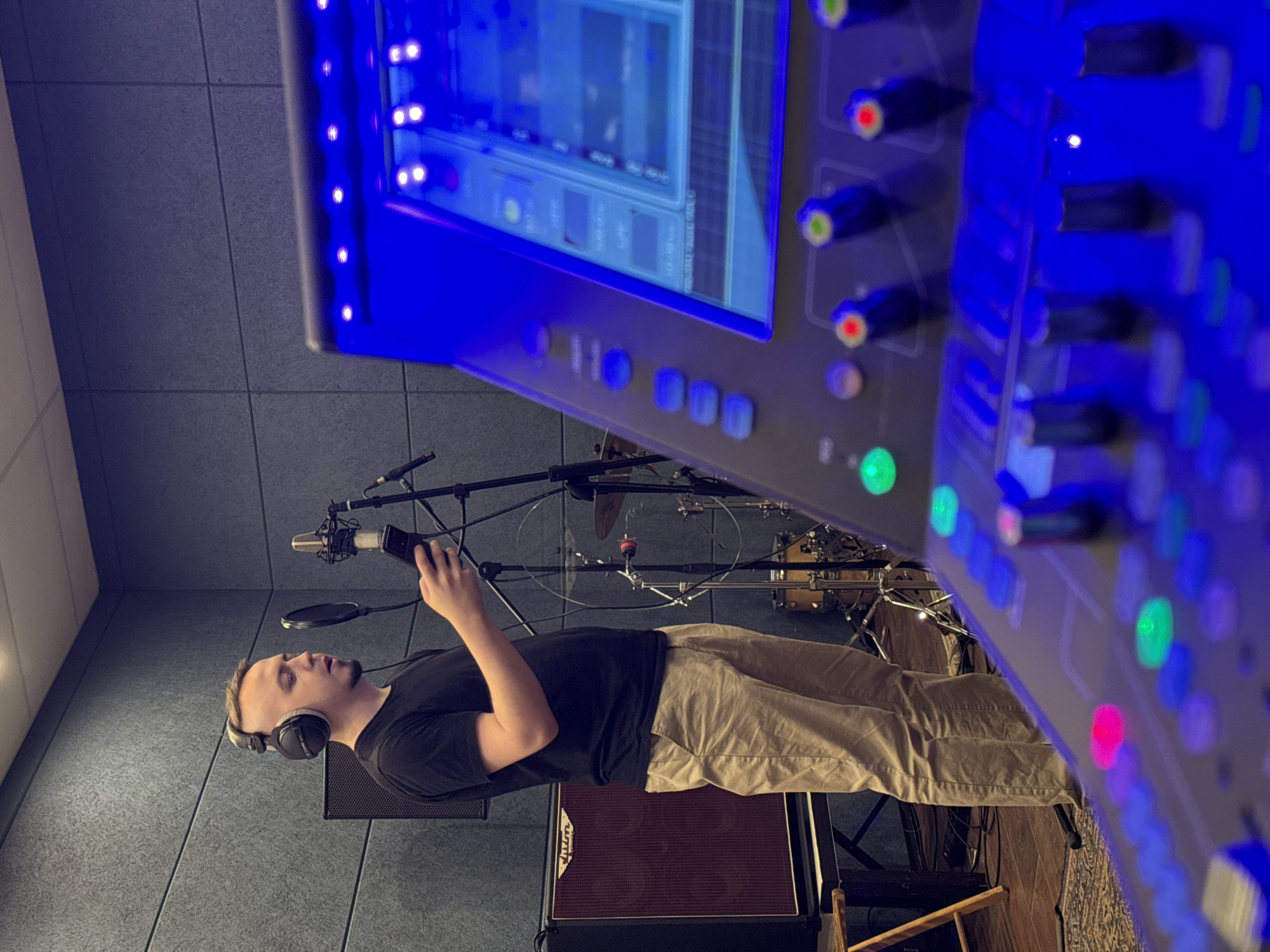 A man stands in a recording studio, wearing headphones and holding a device near a microphone, likely recording vocals. The studio features soundproofing wall panels, a mixing console with illuminated buttons and a screen, musical instruments like drums visible in the background, and amplifiers on the floor. The location is not discernible from the image; hence, there is no indication of being in Moscow, Russia.