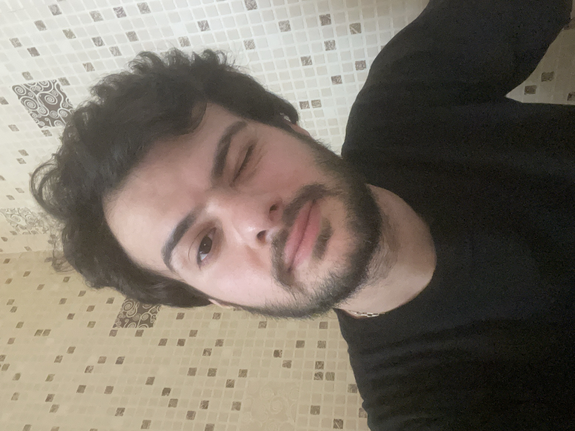 A medium shot photograph captures a young man from the chest up, looking directly at the camera. He has dark, curly hair and a beard. He is wearing a black t-shirt and a silver chain necklace. His right eye is open, while his left eye is closed in a wink. The background consists of a tiled wall with a geometric pattern. The lighting is soft, suggesting an indoor setting. The overall mood of the image is casual and playful. There is no discernible information in the image to suggest the location is Tbilisi, Georgia.