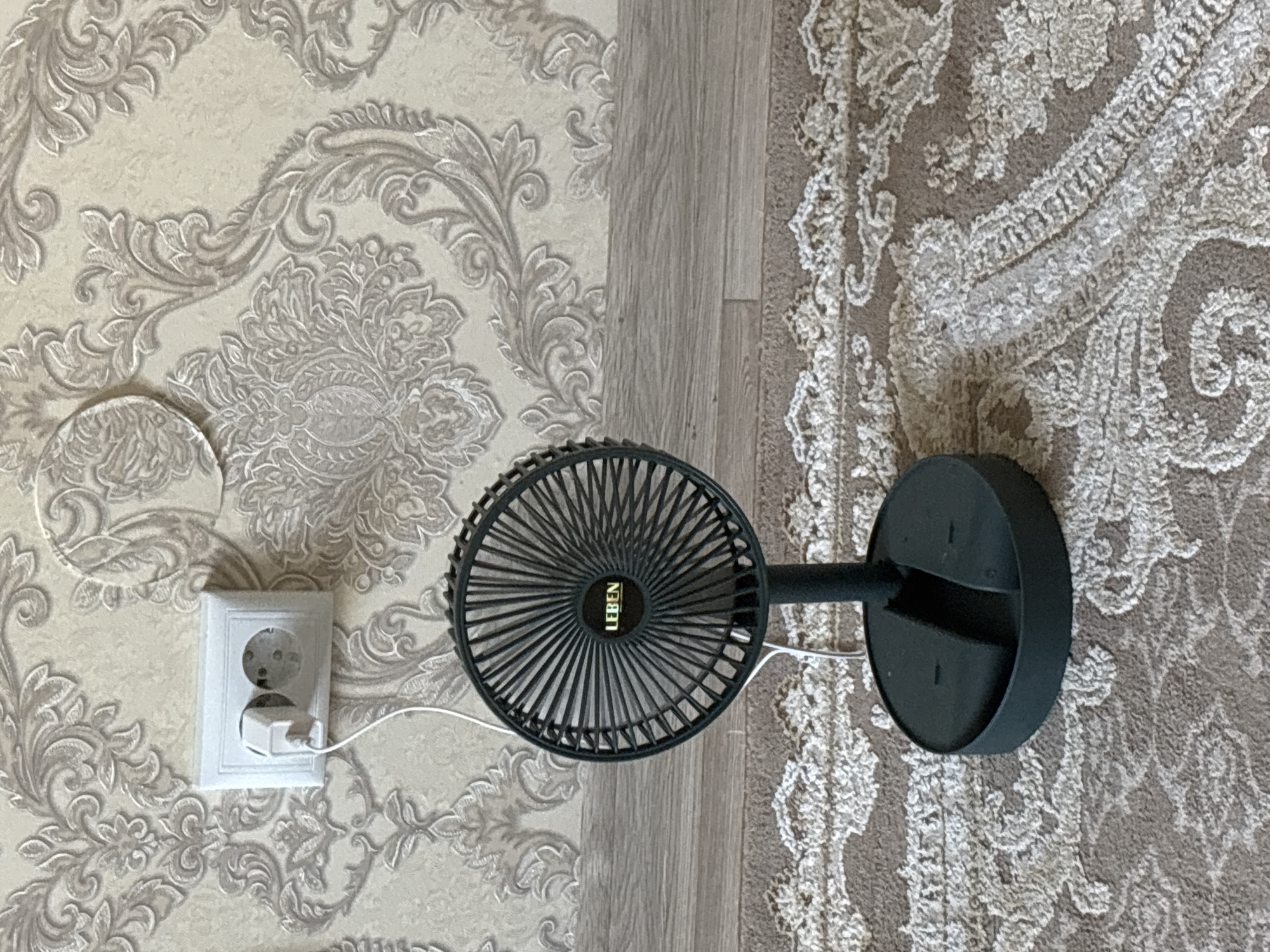 The image depicts a black portable fan with the brand name "LEBEN" visible in the center of its grille, plugged into a white electrical socket on a wall covered with damask wallpaper. The fan is positioned on a floor covered with a patterned rug. The scene is indoors, likely a domestic setting. There are no people or animals visible in the image. The lighting suggests it is daytime, and the environment appears to be a room. The location is not specified in the image, and therefore, cannot be determined to be in Nefteyugansk, Russia.