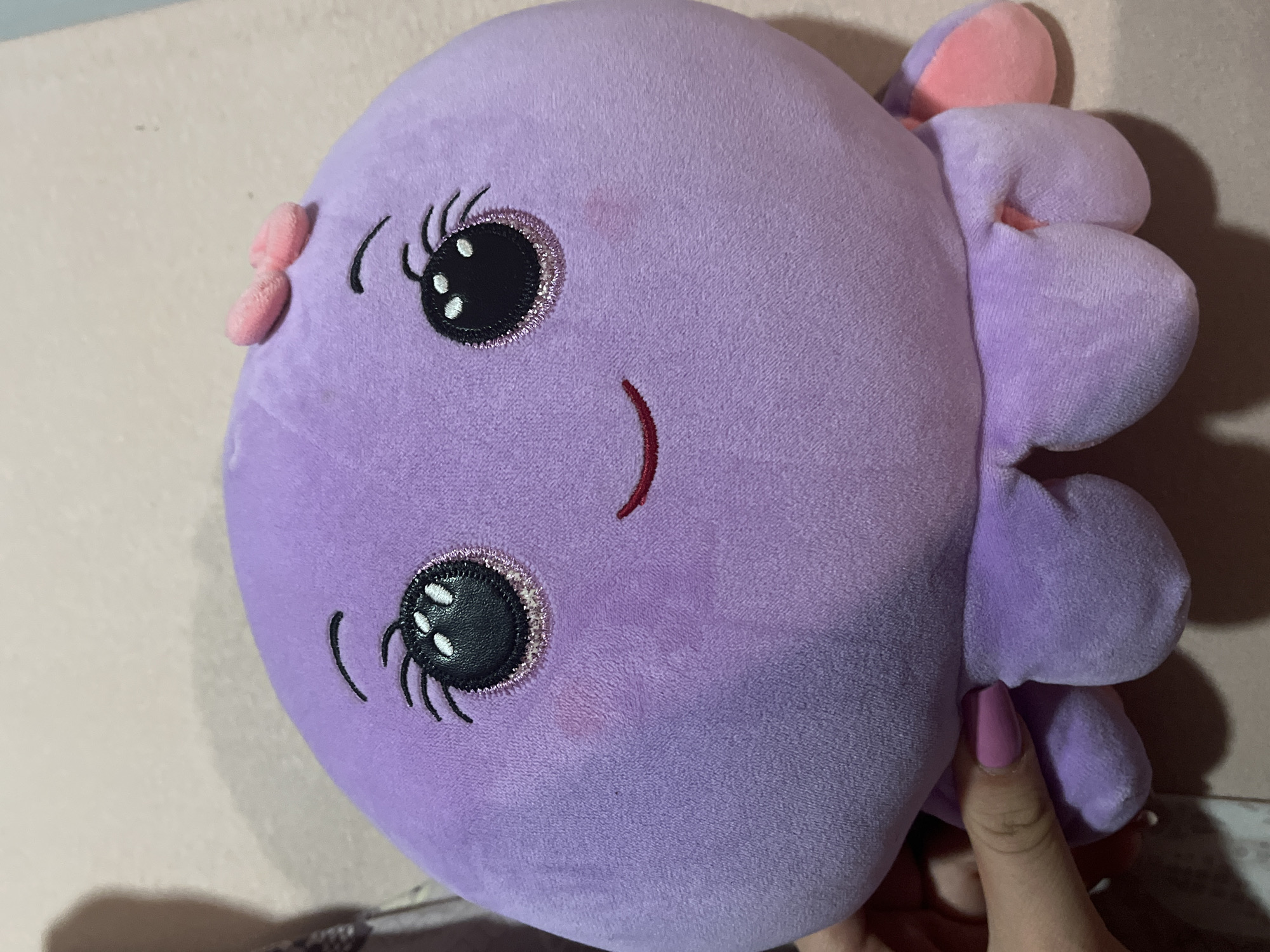 A purple plush octopus toy with pink accents is shown. The toy has large, embroidered eyes with glittery pink accents and black eyelashes, a curved red mouth, and two pink ear-like appendages on its head. It has a textured surface with several bulbous protrusions along its right side. A faint pink stain is visible on the toy's left cheek area. A person's hand with pink nail polish is visible on the right side of the image, holding the toy. The background appears to be a neutral-colored surface, possibly a wall or furniture. There is no visible indication of the city of Plovdiv, Bulgaria in the image.