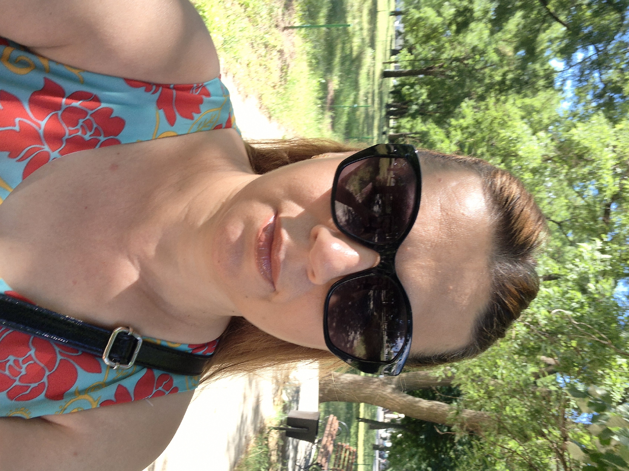 The image shows a close-up, sideways selfie of a woman outdoors. She is wearing sunglasses and a blue and red floral print top. A black strap with a silver buckle, likely from a bag, is visible across her chest. Her hair is brown with lighter streaks and falls over her shoulder. The background is blurred but shows greenery, trees, and sunlight, suggesting it is daytime and the weather is pleasant. The overall impression is casual and relaxed, possibly a selfie taken during an outdoor excursion or while enjoying a sunny day. There is no visible text in the image.