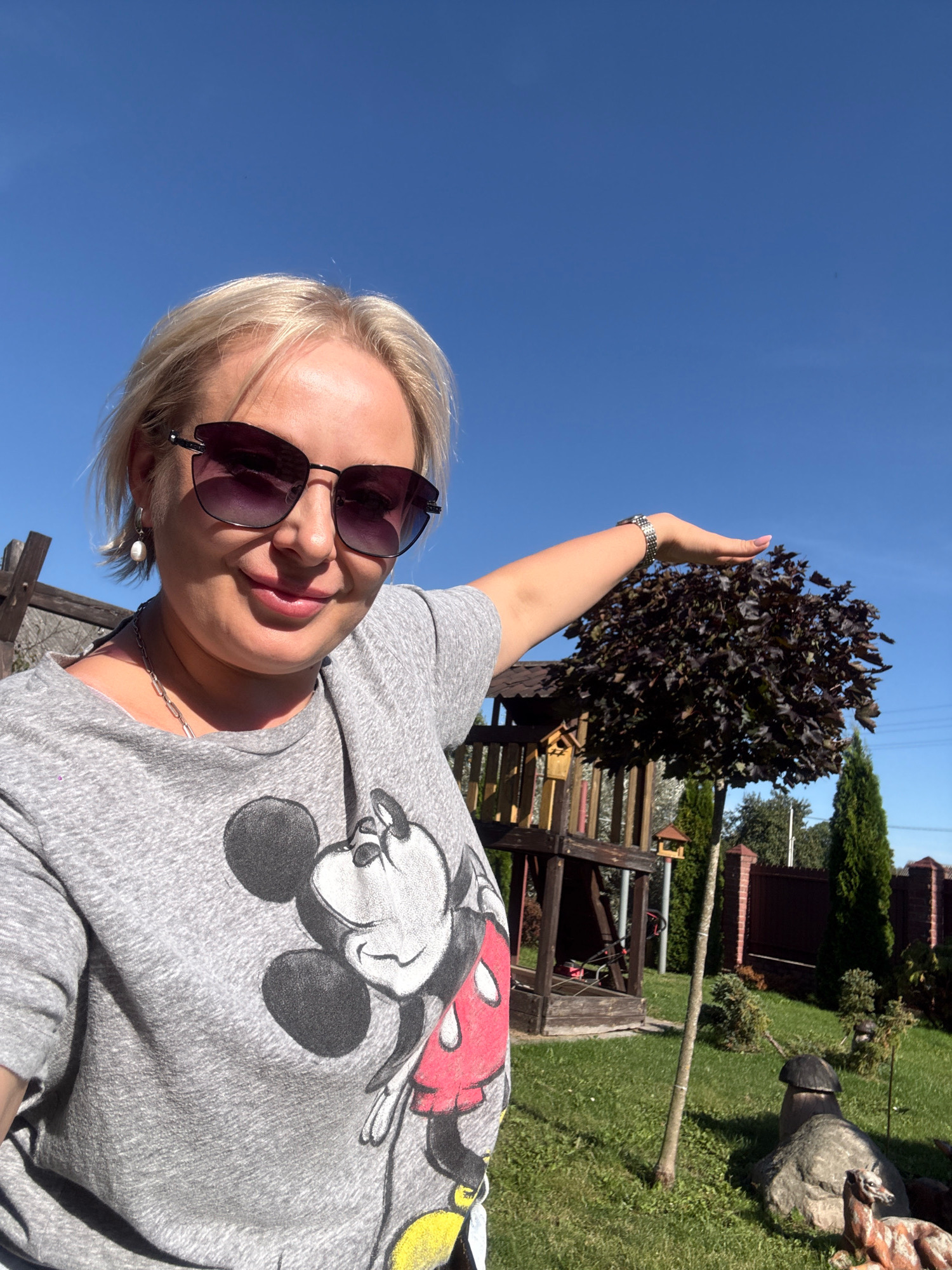 A woman with blonde hair and sunglasses takes a selfie in a backyard. She is wearing a grey t-shirt with a Mickey Mouse graphic. Her left arm is extended, and her hand is positioned above a small, dark-leaved tree. Behind her is a wooden play structure and a brown fence. The sky is clear and blue, indicating a sunny day. The location is the city of Slawharad, Belarus.