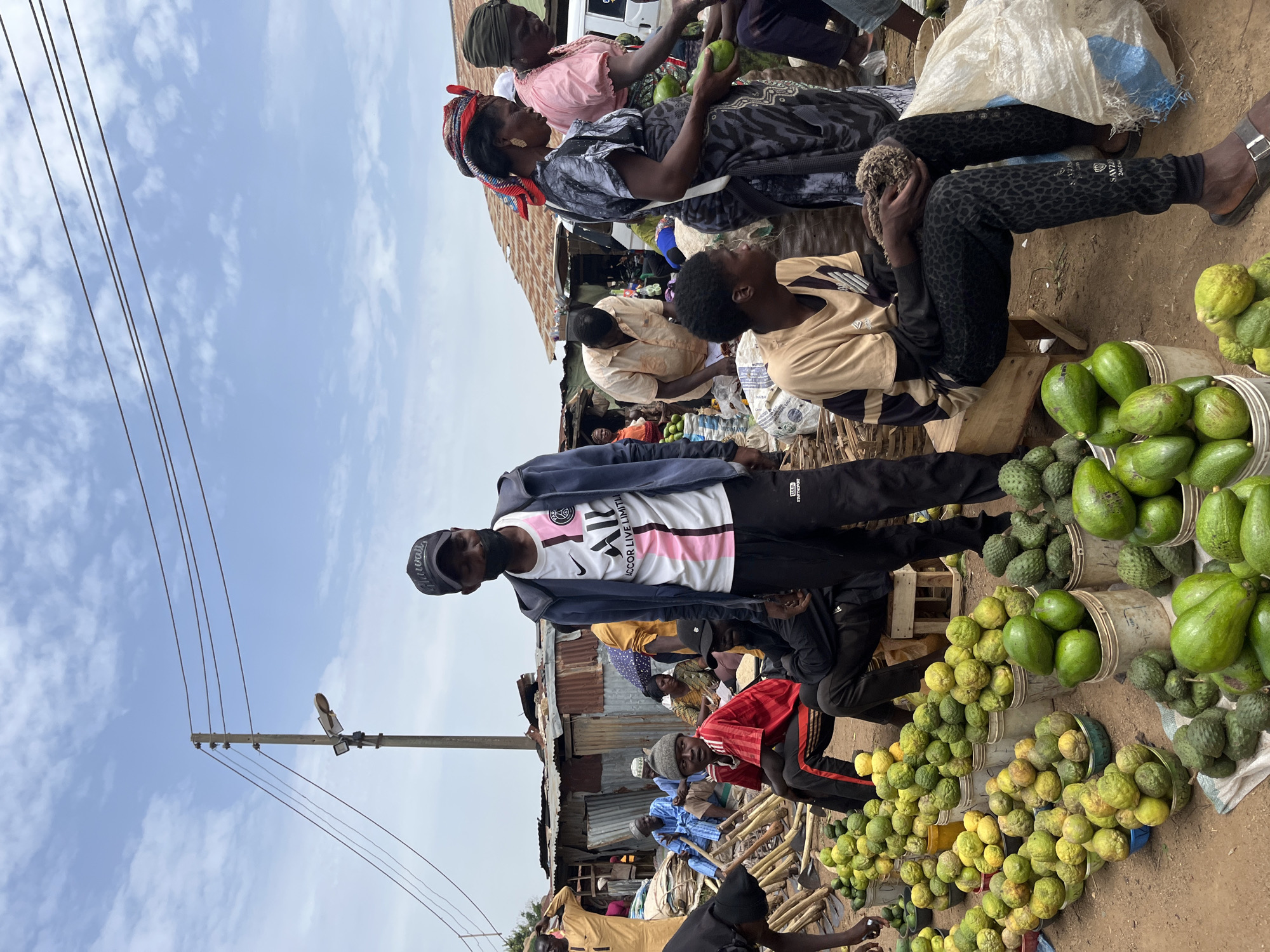 This image depicts a bustling outdoor market scene in Gura Top, Nigeria. The primary subjects are a diverse group of people, including women and men of various ages, engaged in selling and interacting at market stalls. Numerous objects are visible, notably overflowing buckets and baskets filled with a variety of fresh produce. Avocados and other green, bumpy fruits dominate the foreground, with some yellowing fruits also present. The setting appears to be a roadside market, characterized by corrugated iron shacks, wooden structures, and a sandy ground. Overhead, power lines stretch across the frame, and a utility pole with a light fixture is visible.
The activity taking place is clearly commerce – the buying and selling of goods, primarily fresh produce. People are browsing, handling the fruits, and presumably negotiating prices. The atmosphere seems lively and dynamic, with many individuals present and actively participating.
Notable details include the clear, bright daylight, suggesting it's daytime, likely morning or afternoon. The weather appears fair with a blue sky and scattered clouds. The expressions on the faces of the people are varied, some appear focused on their transactions, while others seem more relaxed or observant. The abundance of produce signifies a thriving local agricultural trade.
The image shows some visible text on the clothing of one individual. On a white football jersey with pink and black accents, the text "ACCOR LIVE LIMITI" is legible, along with a Nike logo and "JORDAN" text on a black jacket. Another person's shirt displays "LLOYD" in white text on a black sleeve. The overall scene captures the vibrant essence of a local Nigerian market.