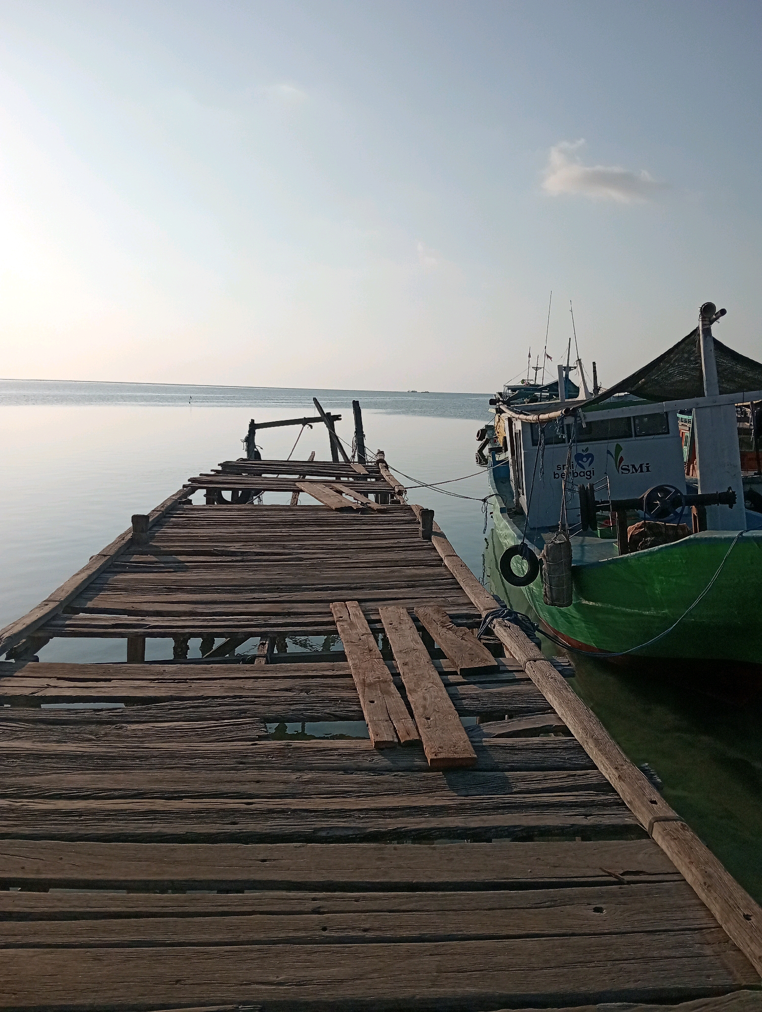 The image depicts a dilapidated wooden pier extending into calm, light blue water under a clear sky. The pier is weathered, with several planks missing and others askew, revealing the water below. To the right of the pier, a green and white fishing boat is docked. The boat has a cabin with windows and various equipment, including fishing rods and a lifebuoy. The water is still, reflecting the pale sky. The scene appears to be at a location in or near Pagerungan Kecil, Indonesia, as suggested by the context. The lighting indicates it is likely daytime, possibly early morning or late afternoon, with soft sunlight. No people are visible in the image. The environment is tranquil and shows signs of natural wear and tear on the man-made structure.