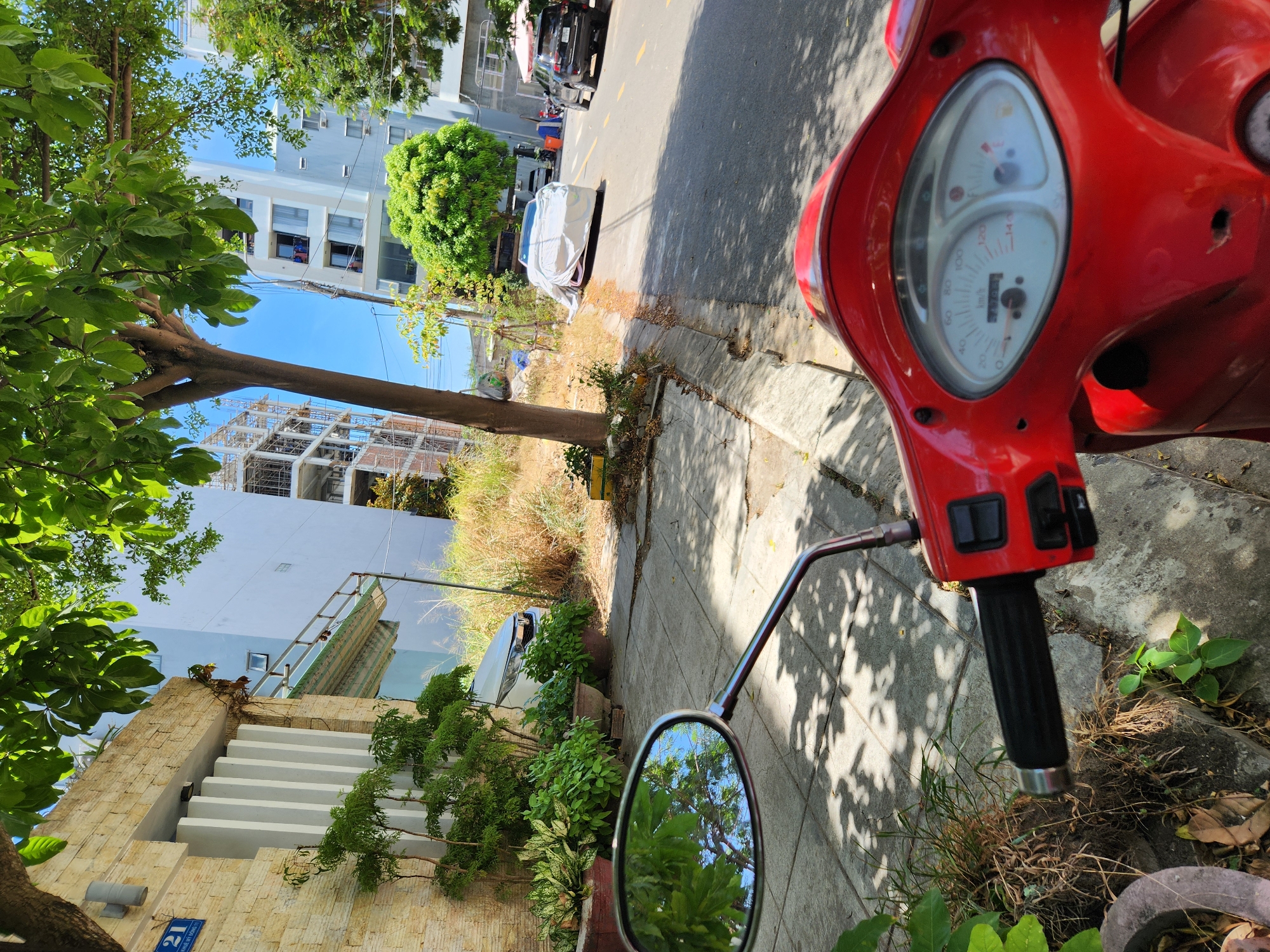 The image depicts a street scene in Da Nang, Vietnam. In the foreground, the red handlebars and dashboard of a scooter are visible, with a rearview mirror showing reflections of green trees and a blue sky. The scooter is parked on a paved sidewalk adjacent to a street.
On the left side of the street, a building with a facade of light-colored stone blocks and white vertical elements is present. A blue sign with the number "21" is affixed to this building. Further down the street, a modern building with light blue walls and multiple windows is under construction. Several trees with lush green foliage are scattered throughout the scene, casting shadows on the ground.
Across the street, a white car is partially covered with a light-colored tarp. Further down the road, a black SUV is parked, and an umbrella with a pink canopy is visible behind it. The environment appears to be an urban setting on a sunny day. The perspective suggests the photo was taken from the vantage point of the scooter rider.