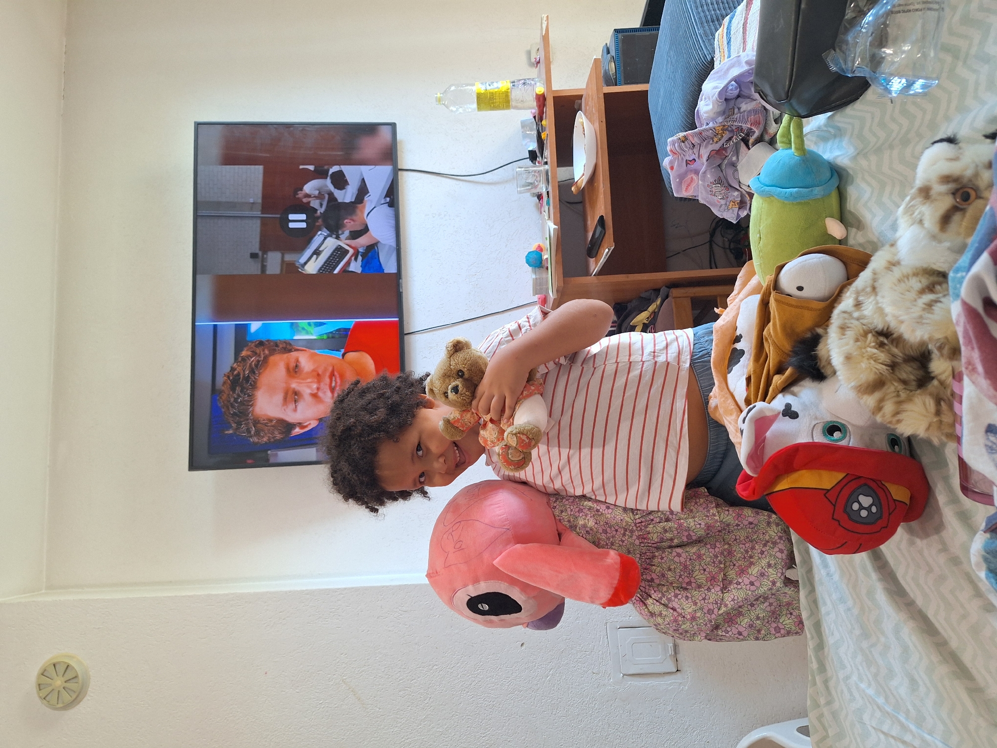 A young child stands on a bed, holding two stuffed animals, a pink flamingo and a small brown bear. The child is wearing a striped t-shirt and shorts. A large television is mounted on the wall behind the child, displaying images of people. A wooden desk with various items is visible to the right of the child, including a bottle of water and a glass. The room appears to be a bedroom, with a bed and other personal belongings. The scene is illuminated by natural light. The location is identified as Blagoevgrad, Bulgaria.
