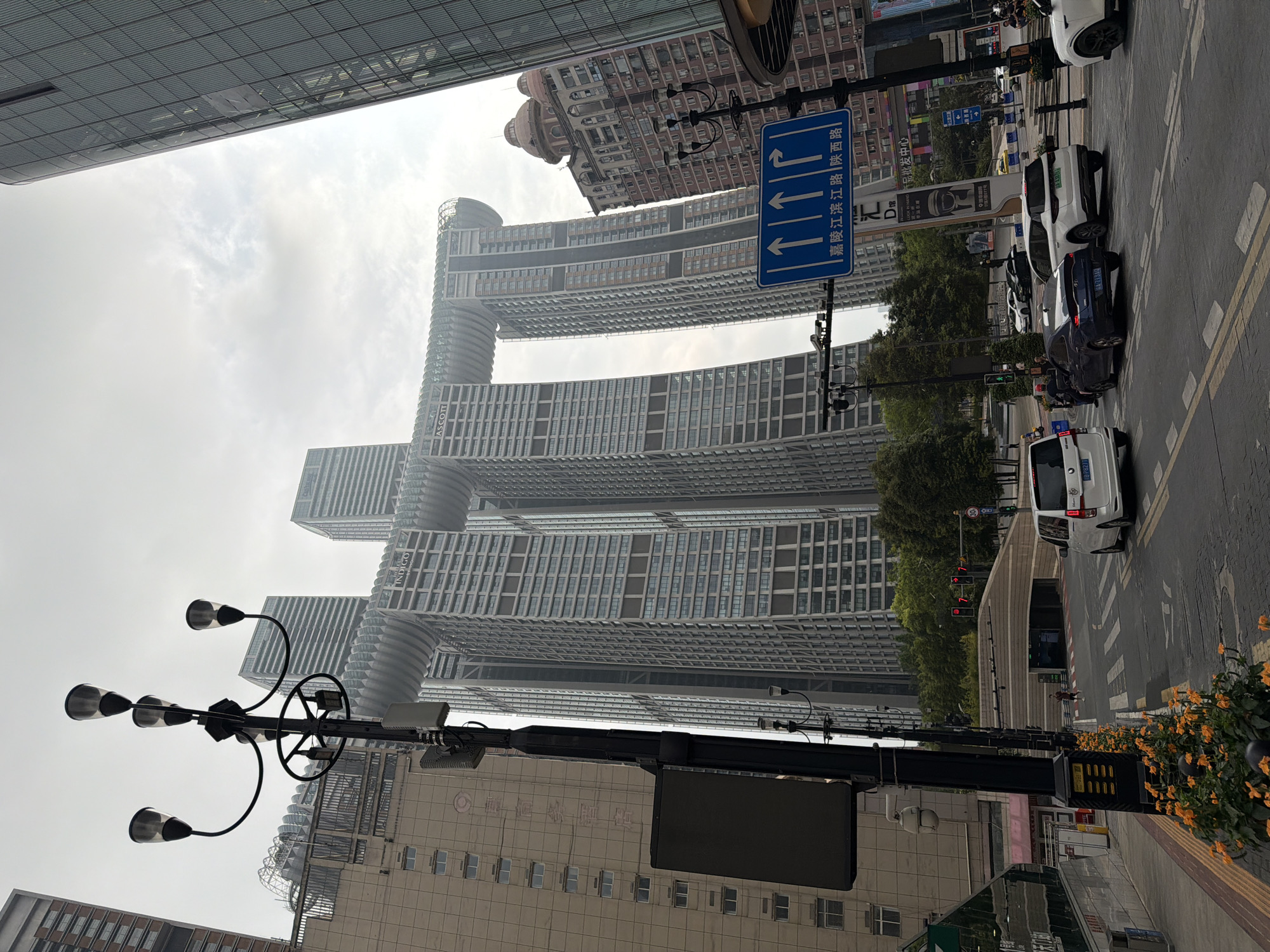 The image shows a street scene in Yuzhong District, China, dominated by modern architecture. In the foreground, a black lamppost with multiple lights stands to the left, equipped with a digital display and a security camera. Orange flowers are planted along the sidewalk. The street features a crosswalk and several vehicles, including a white minivan, a dark blue sedan, and a white SUV. In the background, the distinctive Raffles Chongqing complex, also known as the "Crystal," is prominent. This architectural marvel consists of three connected skyscrapers with a horizontal structure resembling a ship or a bridge spanning between them. Other tall buildings, including one with a domed roof and a facade adorned with red brick patterns, are also visible. A blue traffic sign with white arrows and Chinese text is placed near the lamppost, indicating directional information. Trees line the street, and the sky is overcast. Pedestrians are sparsely visible on the sidewalks and crossing the street. A tunnel entrance is visible further down the road.
