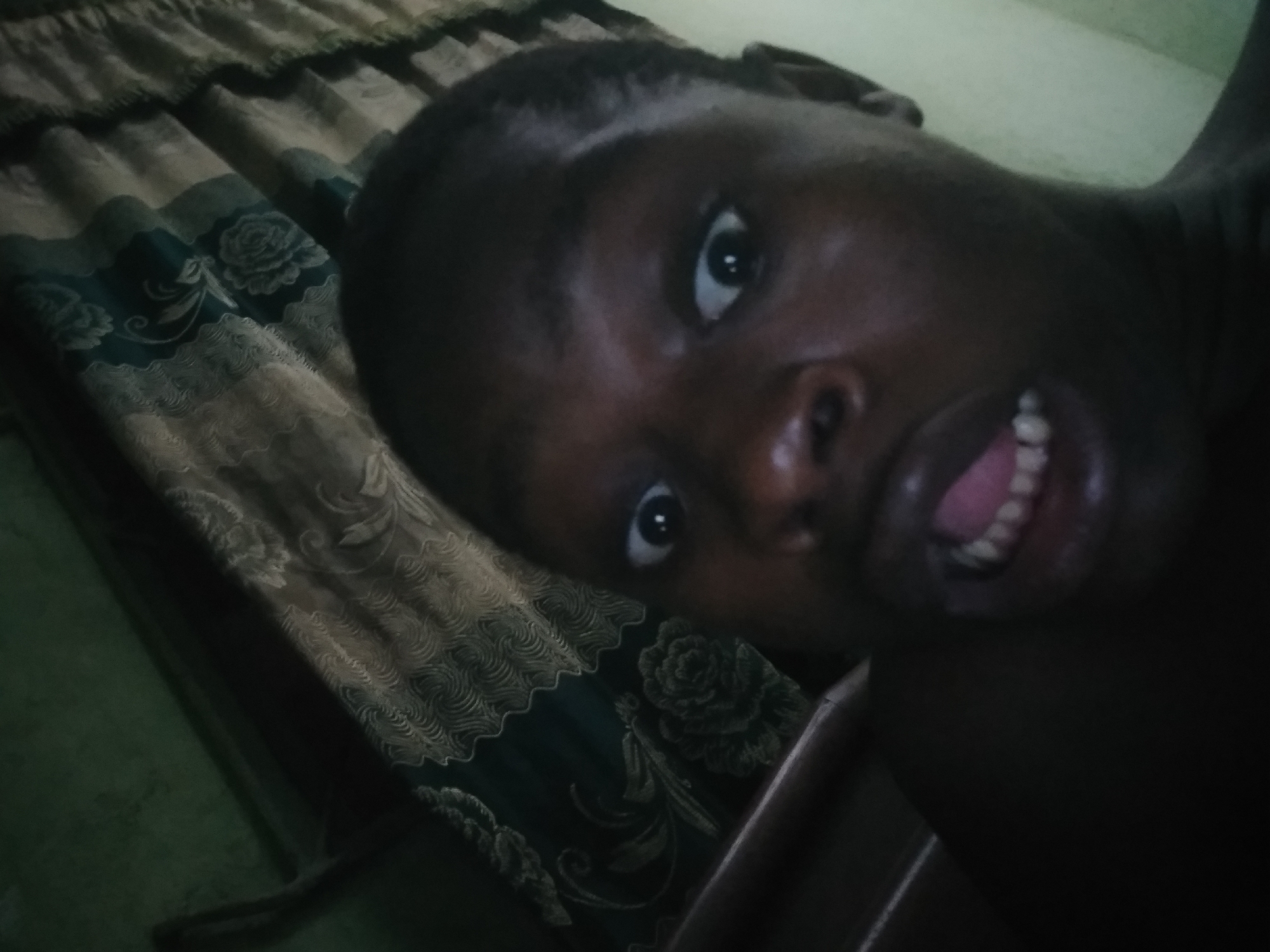A person with dark skin is visible in a selfie-style image. They are looking at the camera with their mouth open, revealing their teeth and tongue. The person is indoors, lying on or next to a surface covered in a patterned, beige and green fabric. Based on the location requested, the image appears to be taken inside a room located in Shangisha, Nigeria. No specific evidence indicates interaction with anyone or specific actions beyond posing for the photo. The environment is dimly lit.