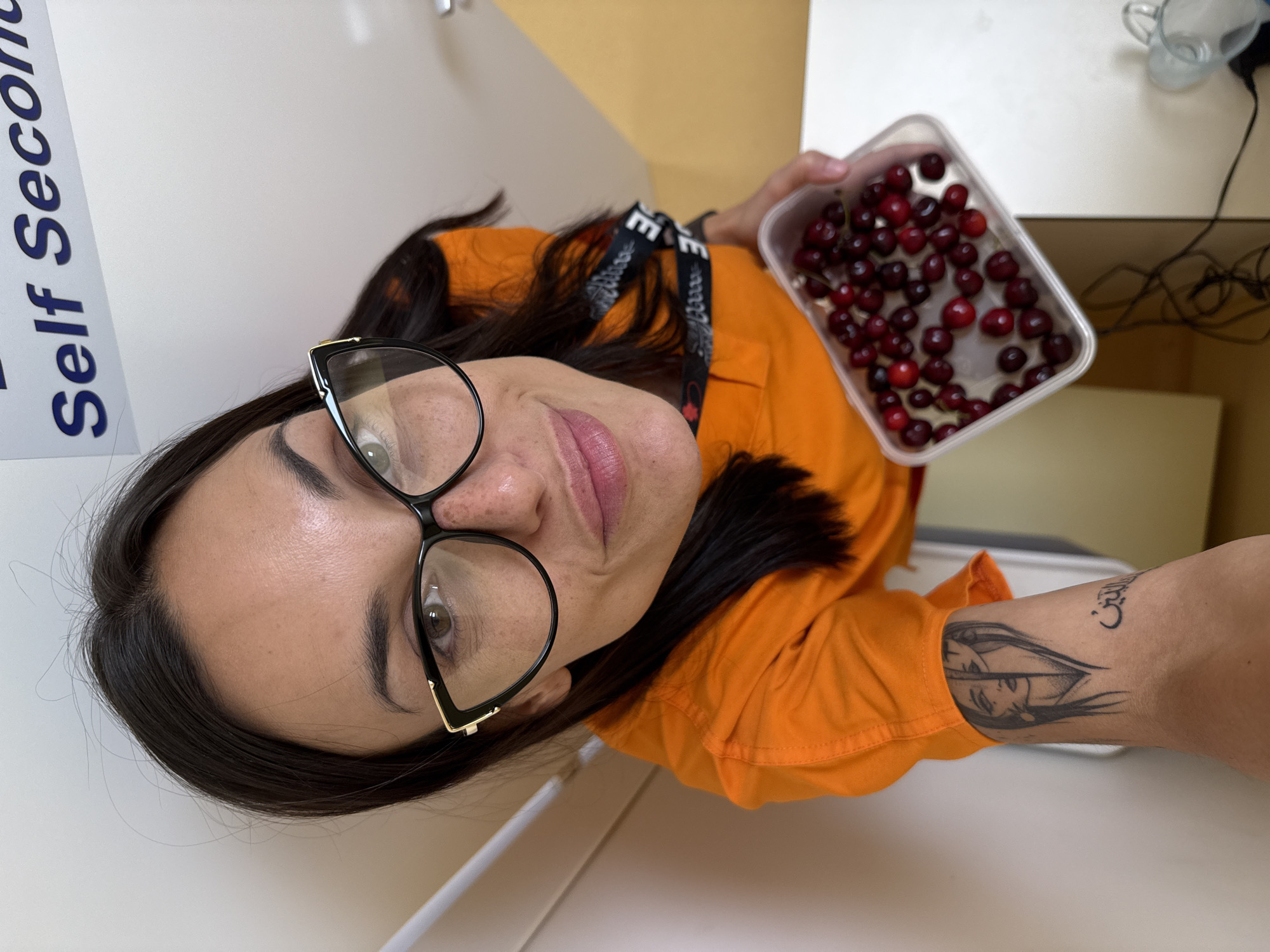 This is a portrait-style photograph of a woman, taken indoors. The woman has dark hair, is wearing large, black-rimmed glasses, and a bright orange shirt. She is holding a clear plastic container filled with cherries. She has a tattoo on her forearm that appears to be a drawing of a face. Visible text on a blue and white sign behind her reads "Self Seco". The overall lighting suggests it could be daytime. The setting appears to be a domestic or office environment, with a white surface and a yellow wall visible in the background. The woman's expression is neutral.