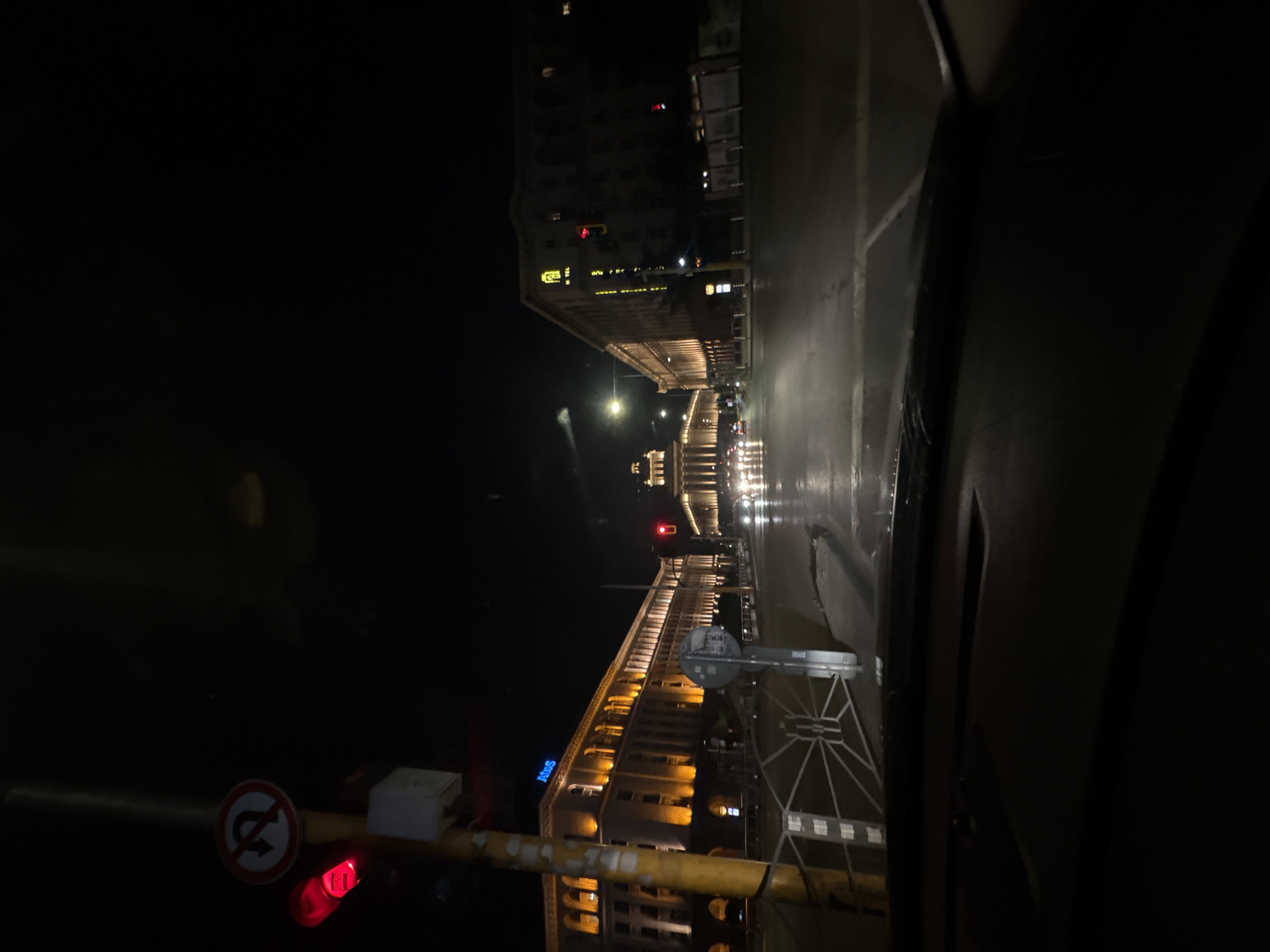 The image shows a night scene in Sofia, Bulgaria. Buildings line the street, illuminated by lights. The Hilton Hotel is visible on the left. Traffic lights are red. The street appears wet, reflecting the lights. A 'No U-Turn' sign is seen. The image is taken from inside a vehicle. The overall scene depicts a typical urban environment at night in Sofia.