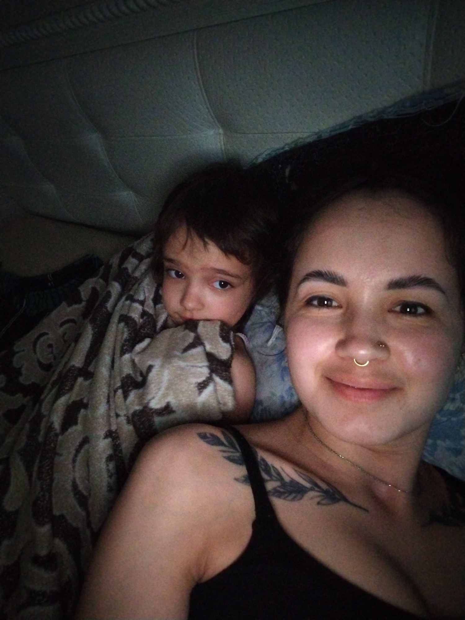 A selfie shows a woman and child in an indoor setting. The woman is on the right side of the frame, facing forward and smiling. She has dark hair pulled back, dark eyebrows, and a nose ring. She wears a black tank top, revealing shoulder and chest tattoos of leaves. A thin necklace is visible around her neck. To the left of the woman, a child is wrapped in a patterned blanket. The child is looking towards the camera. The background includes a tufted headboard and other bedding materials. No elements in this image are related to the city of Noviy, Russia.
