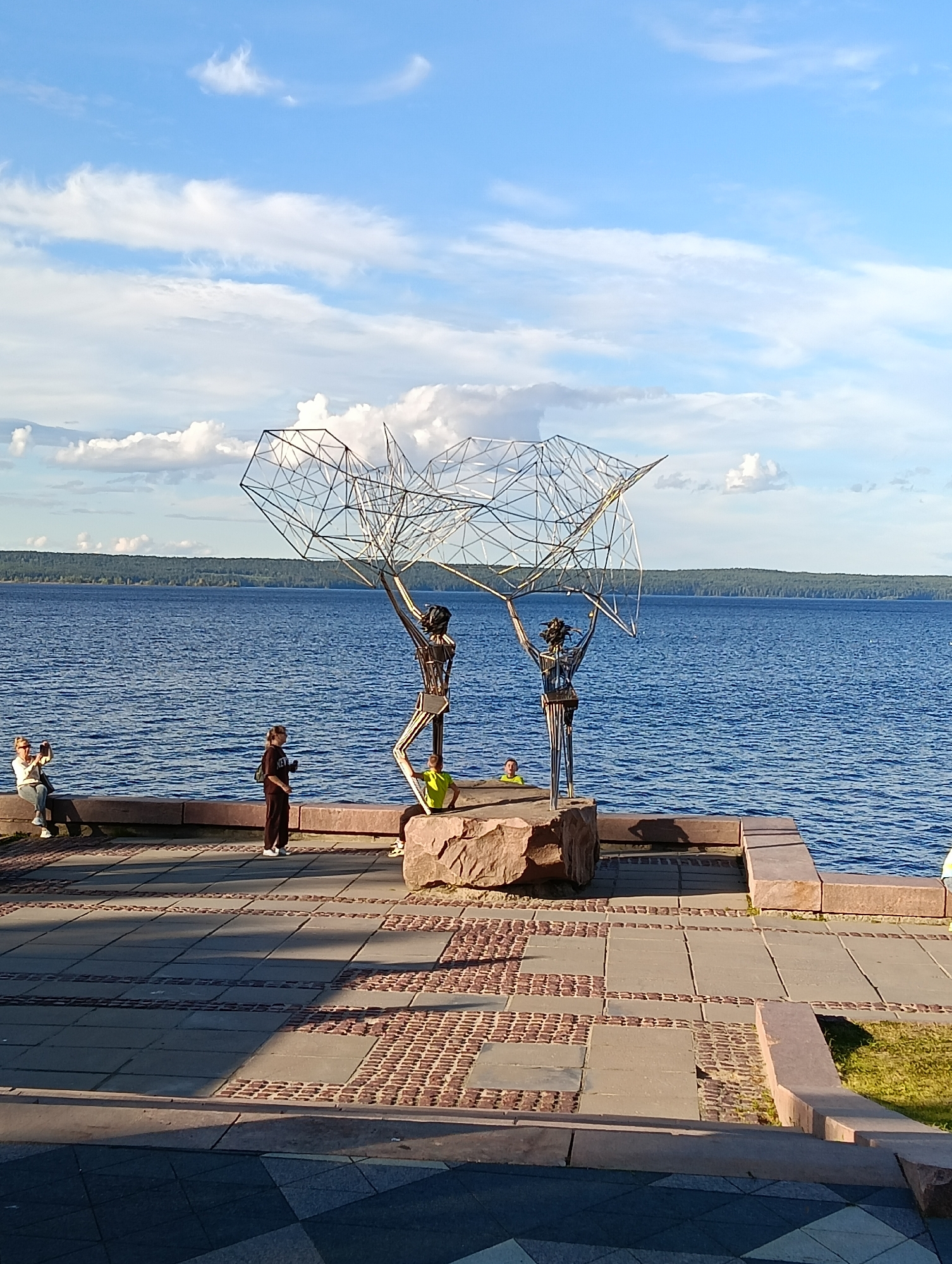 The image depicts a waterfront scene in Petrozavodsk, Russia, likely on the Onega embankment. In the foreground, a patterned paved walkway leads towards the water. To the right, a grassy area borders the walkway. A large, rough-hewn stone sits on the walkway, and a stone bench or wall runs along the edge of the water.
Dominating the mid-ground is a striking metal sculpture of two abstract human figures holding up a large, geometric, wireframe structure. The figures are made of thin metal rods, giving them a skeletal appearance. The sculpture is positioned on a large stone base.
Several people are visible: on the stone wall to the left, a woman in a white top and jeans is sitting and taking a photo with her phone. Next to her, a person in dark clothing stands, facing the sculpture. To the right of the sculpture, a young person in a lime green shirt sits on the stone base, and another person is partially visible in the water near the sculpture.
The background features the vast blue waters of a lake or bay, with a densely forested shoreline visible in the distance under a bright blue sky with scattered white clouds. The overall impression is a public space designed for recreation and enjoyment of the natural scenery, with a prominent artistic element.