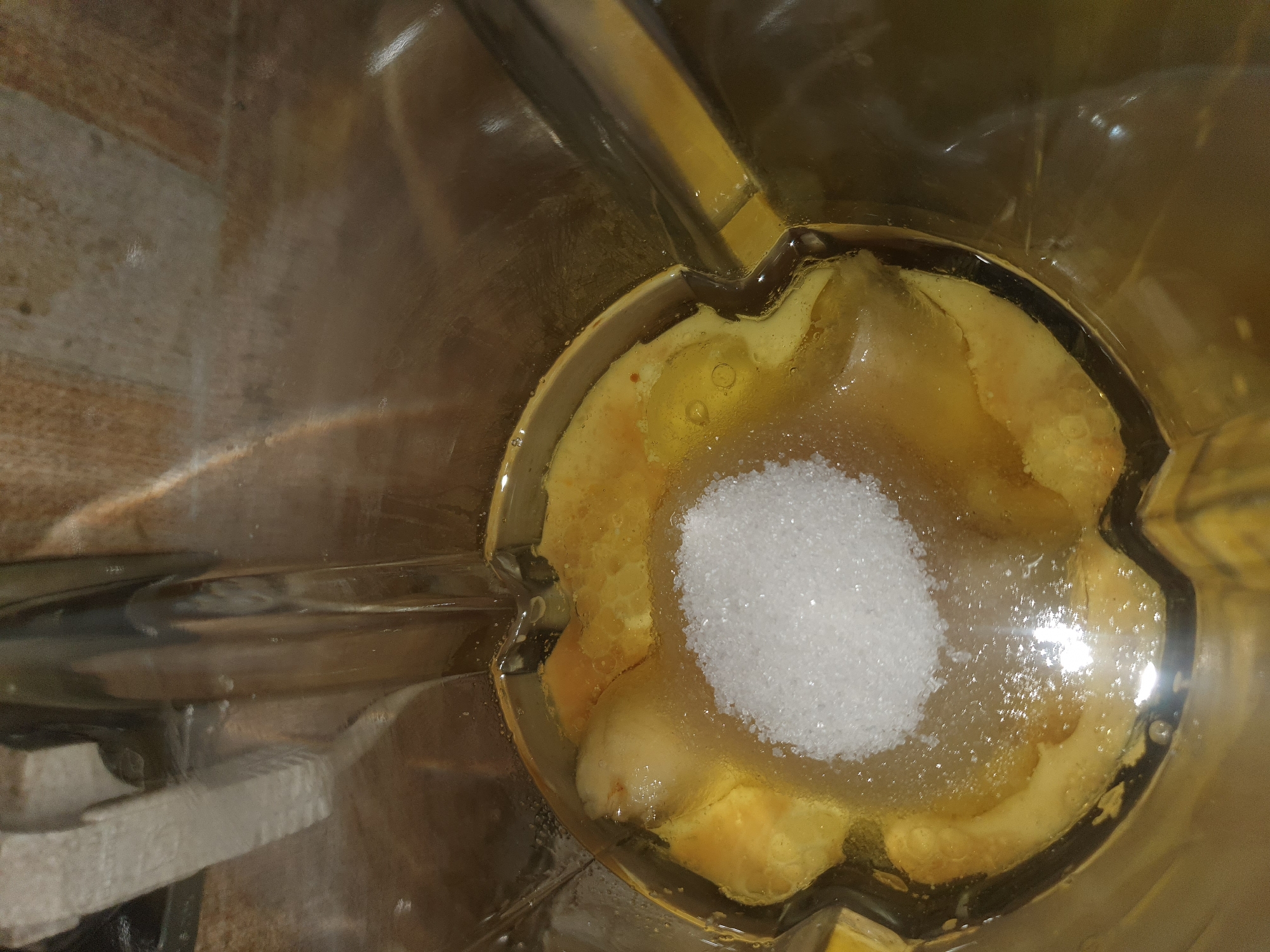 A close-up, overhead shot shows the inside of a blender jar containing several ingredients. A pile of white granulated sugar sits in the center of the frame, on top of a golden-yellow, liquid-like substance. Submerged in the liquid are pieces of what appear to be sliced banana and possibly some other fruit. The blender jar is made of clear plastic and its internal blades are visible at the bottom. The background is out of focus and appears to be a textured surface, possibly a countertop, with warm, earthy tones. There are no people or discernible locations in Shiraz, Iran visible in the image.