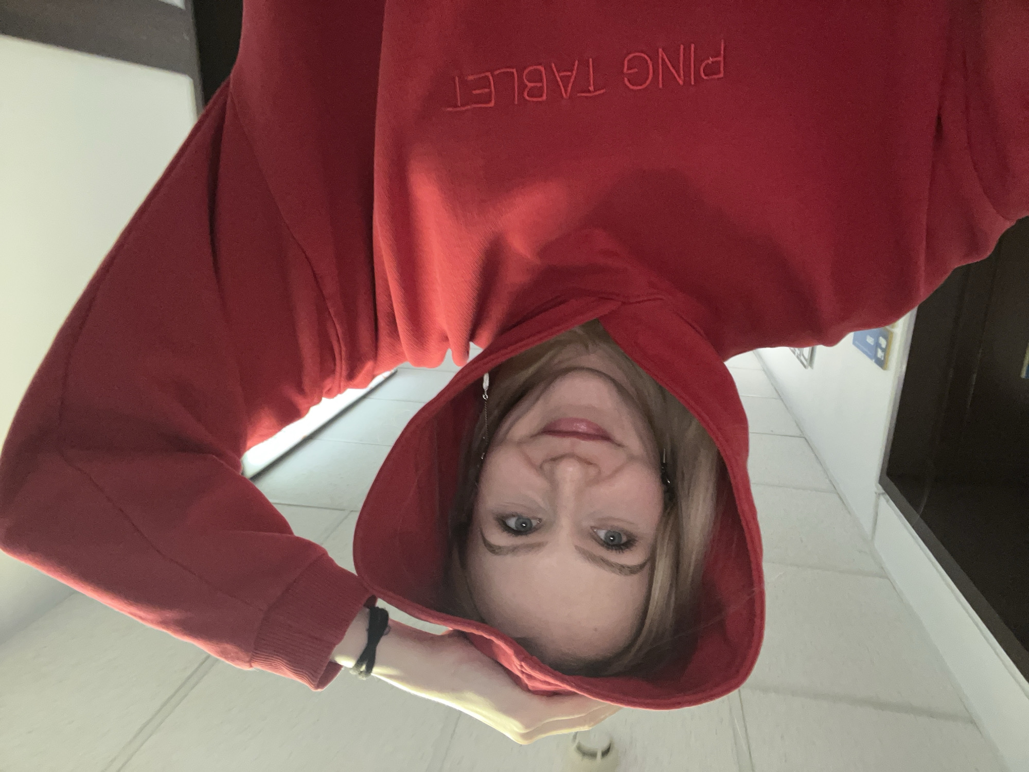 A medium close-up, low-angle photograph captures a woman indoors, presented in an inverted orientation. She is wearing a vibrant red hooded sweatshirt with the hood pulled over her head. Her face, with blonde hair peeking out, is framed by the hood, revealing blue eyes and a gentle, closed-mouth smile as she looks directly at the camera. Visible accessories include dangling metallic earrings and a black wristband on her left wrist, which is positioned near her chin, seemingly holding the hood. Embroidered text across the chest of the red hoodie reads "PING TABLET" when viewed upright. The background is simple, consisting of light-colored walls or a ceiling, with a dark rectangular element on the right side, suggesting an indoor environment. The lighting indicates an interior setting. The overall mood is casual and friendly. This scene takes place in Khimki, Russia.