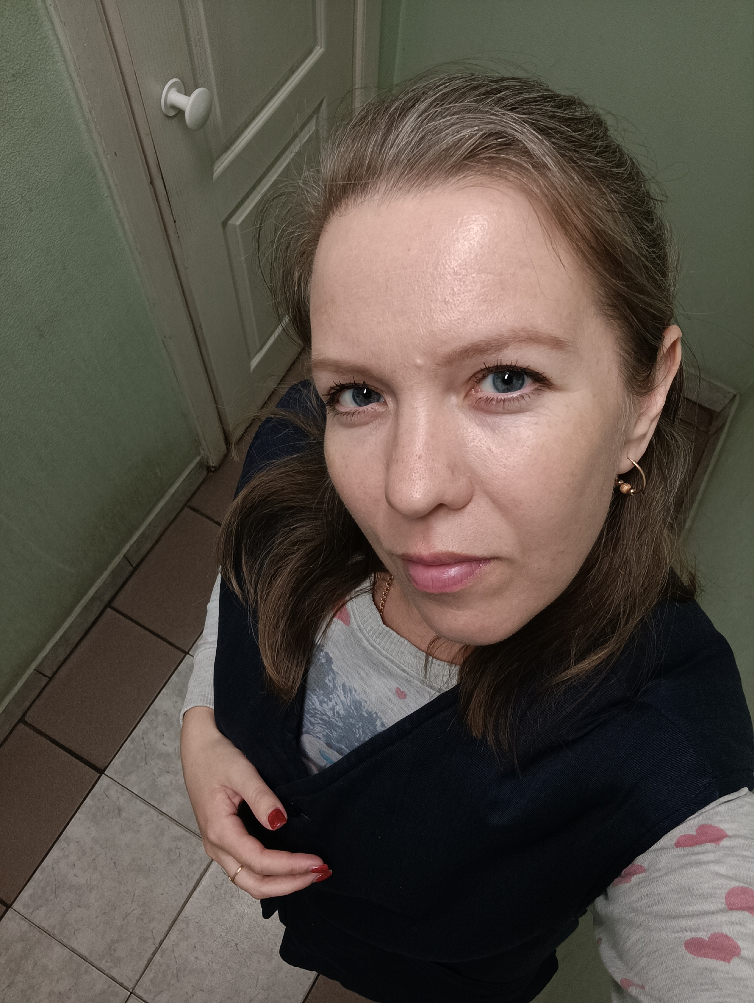 A woman with light skin, blue eyes, and light brown hair with some gray strands visible near the hairline is looking directly at the camera with a slight frown. She is wearing a light gray long-sleeved shirt with pink heart patterns under a dark blue vest or sweater. She has gold hoop earrings and a ring on her left hand with red nail polish.
The background includes a light green wall with a white door and knob to the left. The floor is tiled in alternating shades of brown and gray.
There is no information in the image to indicate this image was taken in Chaikovsky, Russia.