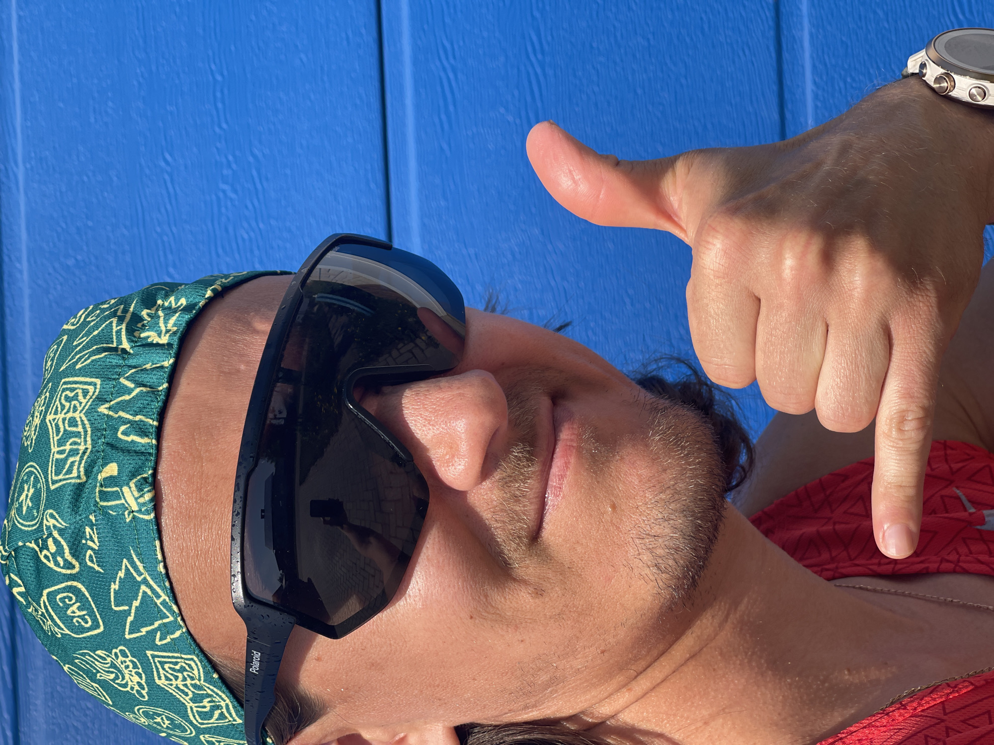 A close-up, eye-level shot features a man with a patterned bandana, dark sunglasses, and a slight beard. He points downward with his right hand, on which he wears a wristwatch. The background consists of a blue, textured surface. The location in the city of Перевозное, Russia, cannot be determined from the visual information provided.