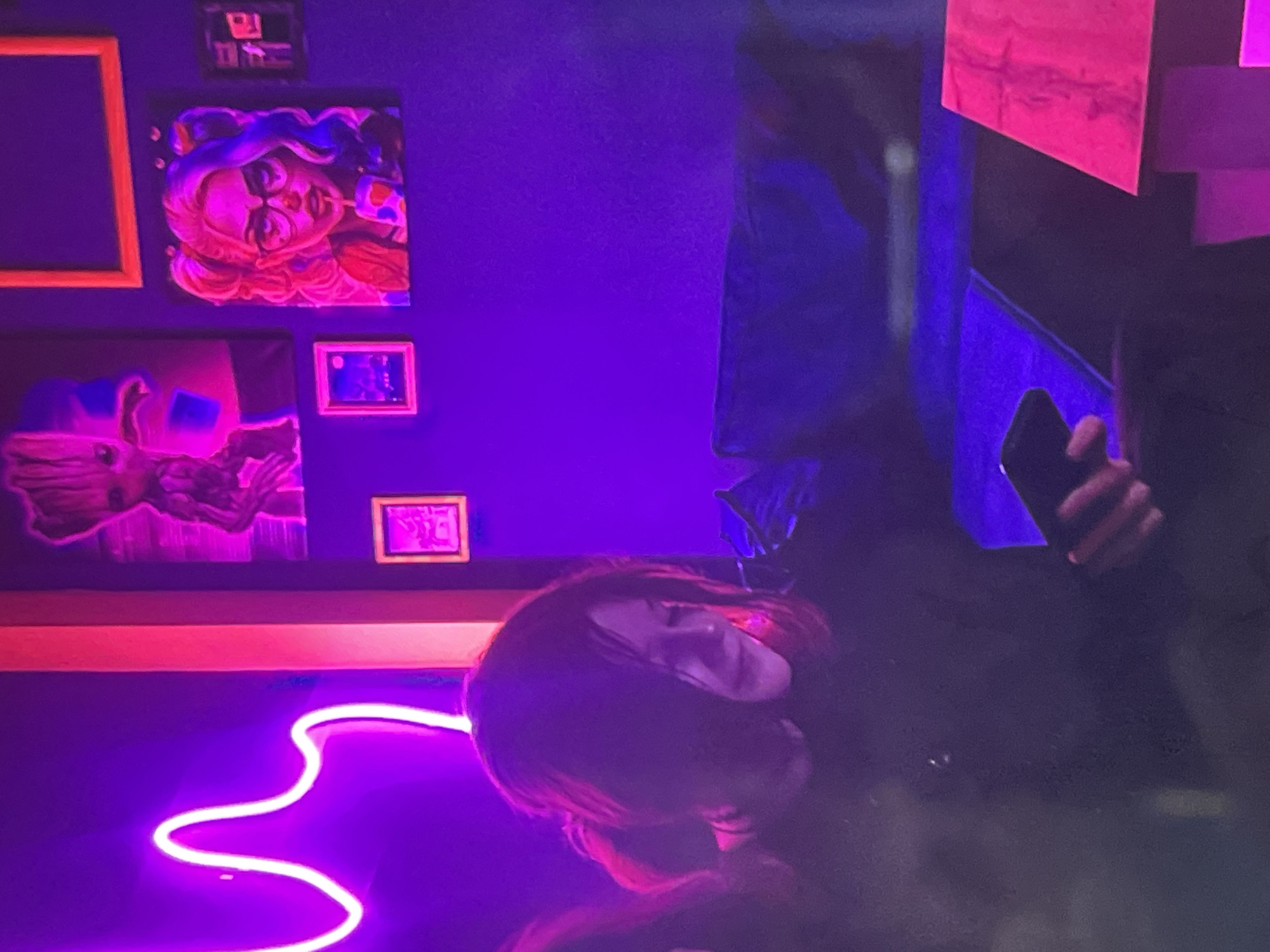 The image depicts a dimly lit indoor scene, likely a cafe or lounge, with vibrant purple and pink neon lighting illuminating the space. In the foreground, a person with dark hair styled in pigtails is seated, looking down at a smartphone they are holding. They are wearing a dark top. The background features a wall adorned with artwork, including a picture of Groot from Guardians of the Galaxy and a portrait of a woman with colorful hair, possibly Harley Quinn. Small framed pictures are also visible on the wall. A dark couch is present in the background, and a table with a pink surface is situated in the lower right corner. The overall ambiance suggests a modern, possibly themed, entertainment venue. The scene is set in Minsk, Belarus.