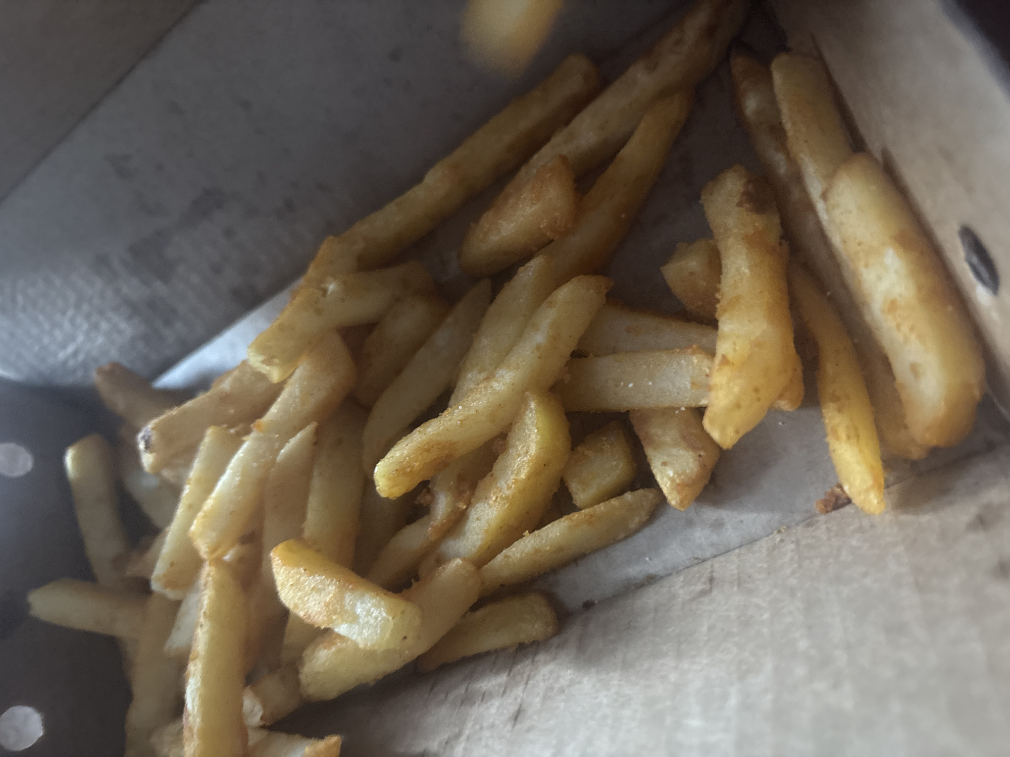 A close-up, overhead shot shows a portion of french fries in a brown cardboard box, likely a takeout container. The fries are golden brown and appear to be seasoned with a light orange spice. Some fries are overlapping and piled on top of each other. The lighting is dim, with some areas of the fries appearing brighter than others. The background is a blurred view of the interior of the box, which is also made of brown cardboard. There is no indication of the city of Aktobe, Kazakhstan in the image.