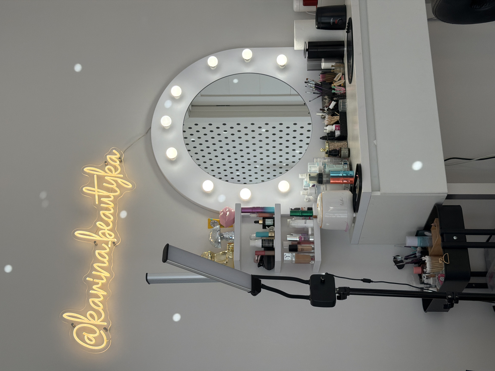 The image shows a vanity area, likely in Riga, Latvia, based on the text visible in the image. A white vanity table is set against a gray wall, illuminated by a bright white vanity mirror surrounded by light bulbs. A yellow neon sign that reads "@karinabeautyka" is attached to the wall to the left of the mirror.
The vanity is organized with various beauty products, including makeup brushes, cosmetics, and skincare items. A small white organizer holds multiple makeup tubes, and other containers of creams and liquids are organized on the vanity surface. A black trolley cart is positioned to the right of the vanity, holding additional cosmetic items. A rectangular light is mounted on a black stand and is positioned in front of the vanity.
The room has an indoor environment with a well-lit area, and the arrangement suggests a space dedicated to beauty and makeup application.