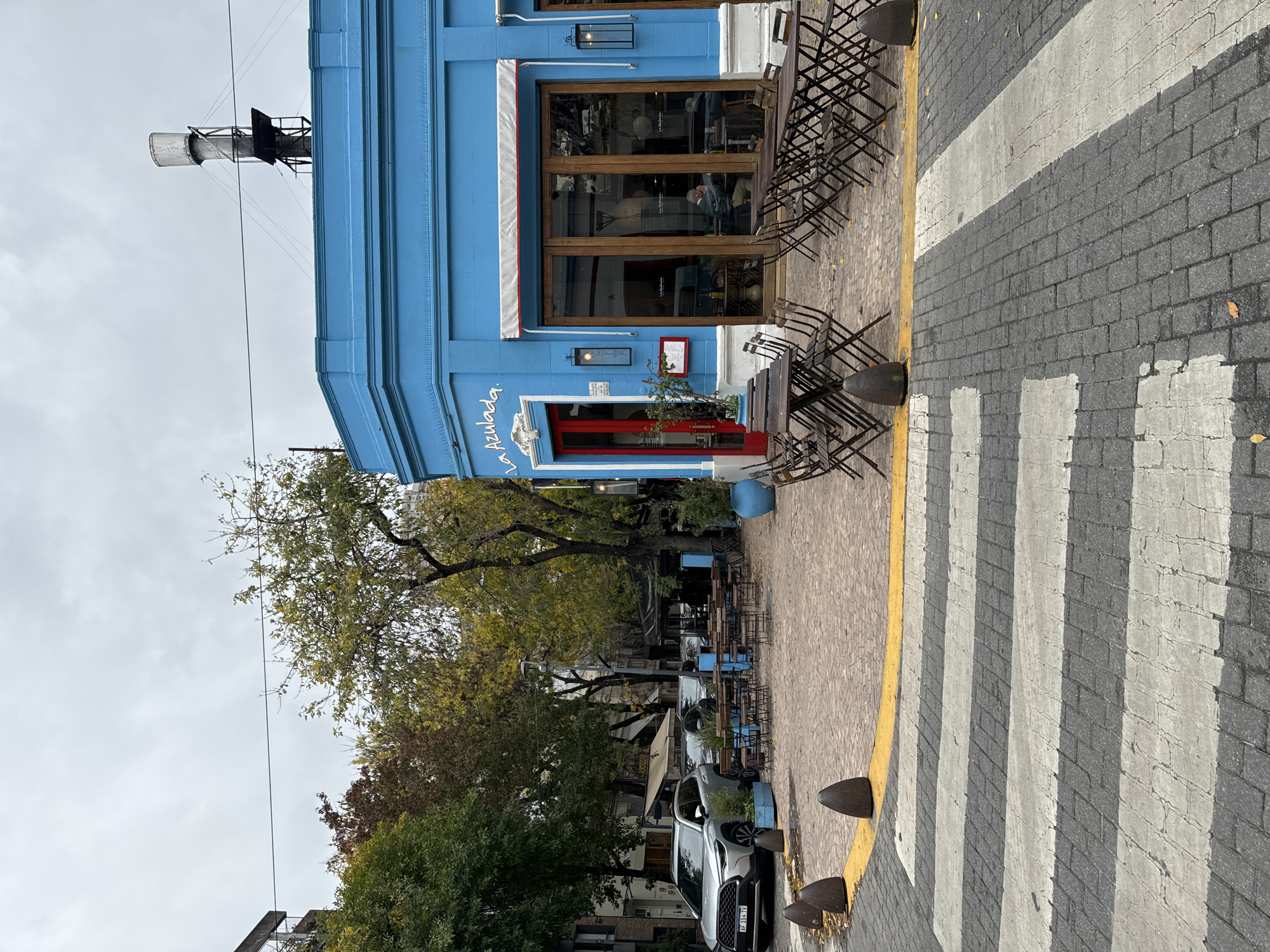 The image shows an eye-level, medium shot of a street in Buenos Aires, Argentina. The prominent feature is a bright blue building with the name "La Azulada" written in white script above a red door. The building has a large window on the right side, and there are several dark-colored metal chairs and tables arranged outside. To the left of the building, there are trees with autumn foliage, some of which obscures additional tables. A silver car is parked along the street. In the background are additional buildings and street furniture. In the upper left corner, there is a metallic structure with a cylindrical top, possibly part of an industrial facility. A crosswalk with white stripes and a yellow line are visible on the right side of the image. The sky is overcast.