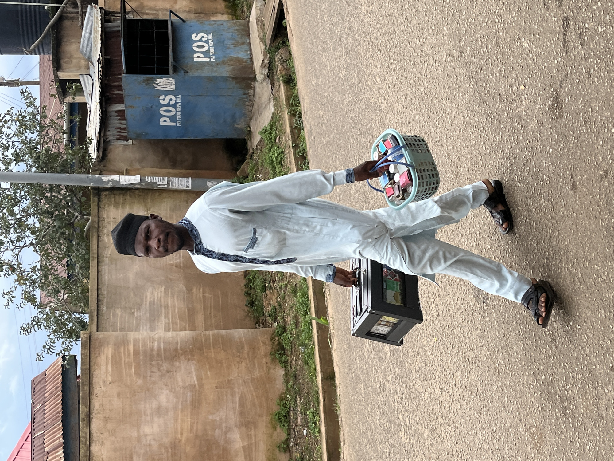 A man is walking on a paved road in Jos, Nigeria, carrying a black display case in his left hand and a light blue plastic basket filled with various items in his right hand. The man is wearing a light blue traditional garment with a black cap. The display case appears to contain several rows of small, colorful items. The basket contains multiple small bottles and boxes, some with labels. Behind the man, a beige concrete wall with a reddish corrugated metal roof structure on top runs along the side of the road. Further down the road, a blue metal structure with "POS PAY YOUR NEPA BILL" visible on it is located. The environment appears to be an urban or semi-urban area with some greenery and overhead utility wires.