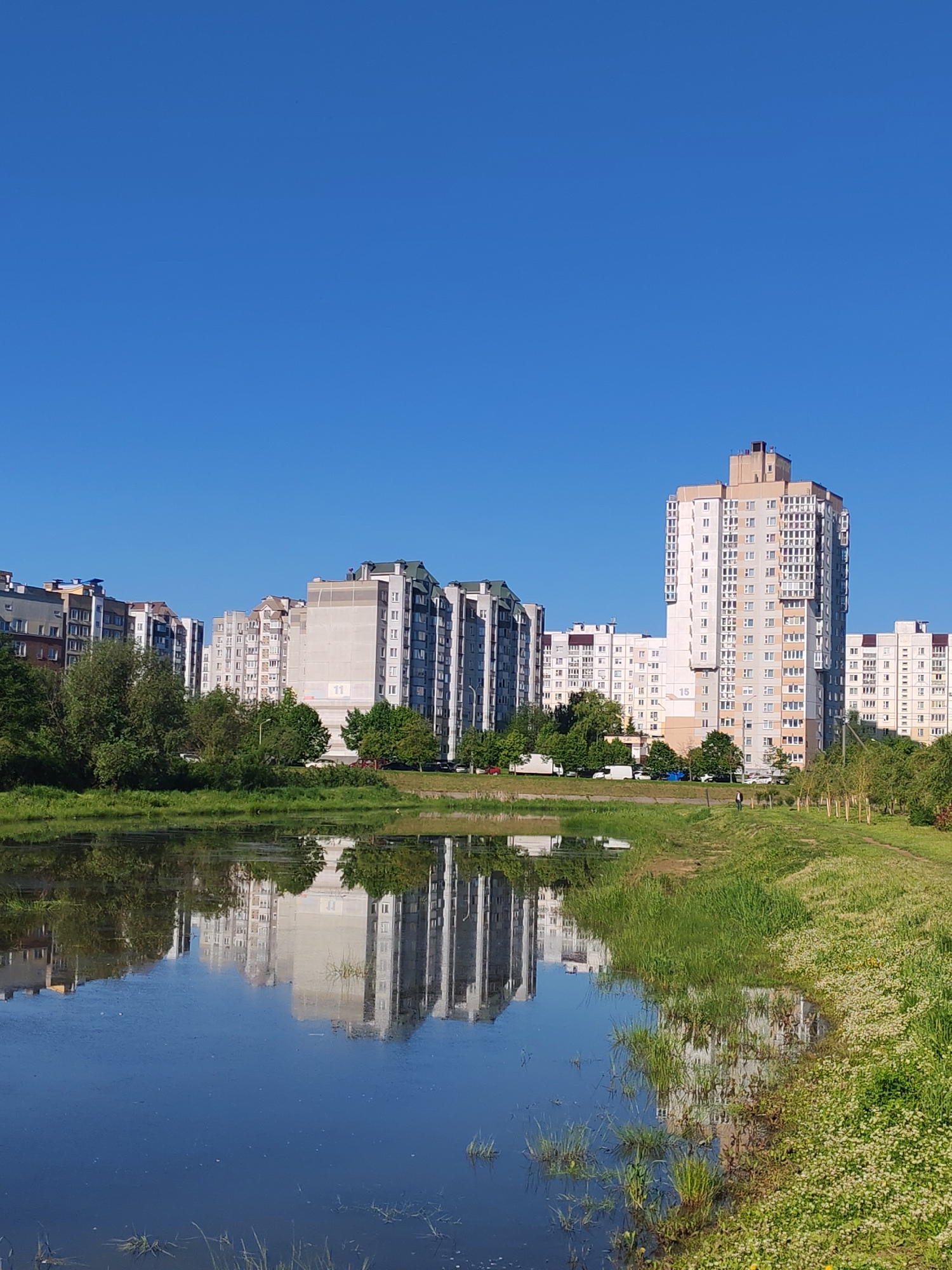 The image depicts a still body of water reflecting a row of apartment buildings under a clear, bright blue sky. The buildings vary in height and color, with some appearing grey and others white or beige. Vegetation, including trees and grass, lines the edge of the water. A solitary figure is walking along a grassy path that runs parallel to the water's edge. The location is in the city of Unknown, Unknown.