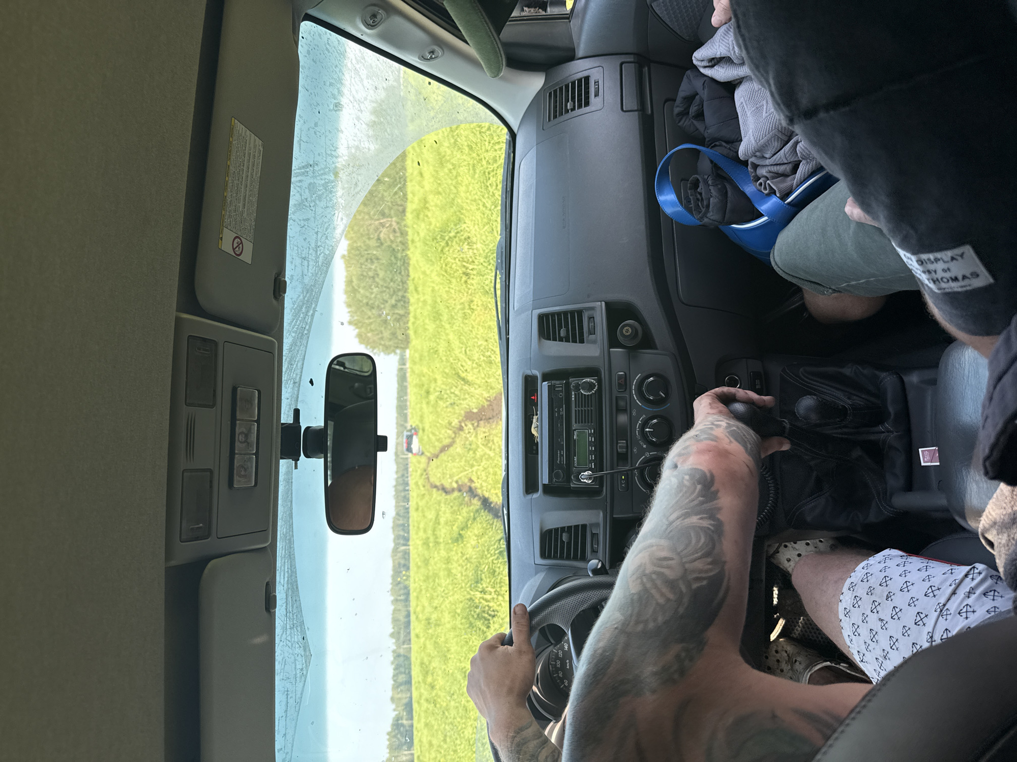 The image depicts the interior of a vehicle, viewed from the perspective of a passenger. The driver's hands are visible on the steering wheel, and their tattooed arm is prominent. The dashboard features a car stereo and climate controls. Through the windshield, a bright green field stretches towards a treeline under a clear sky. A dirt track leads into the field, with another vehicle visible in the distance. The interior of the car contains various items, including clothing, a blue bag, and personal effects. The scene suggests an off-road excursion in a rural or natural environment. The specific location in Malinovka, Russia, is not directly ascertainable from the visual information alone.