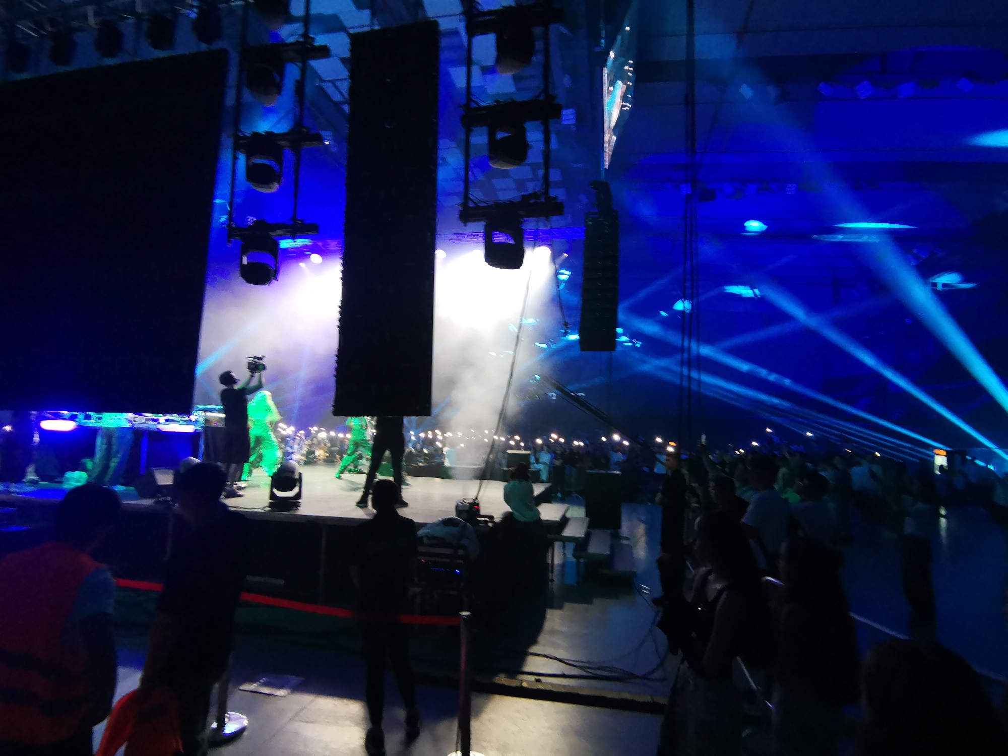 This image captures an indoor live event, likely a concert or performance, within a large venue in Plovdiv, Bulgaria. The scene is primarily dark, illuminated by artificial blue and white lighting, with a pervasive stage haze.
On the left, a large, dark rectangular screen or backdrop is positioned vertically. Above the stage, multiple lighting fixtures, including moving head lights and PAR cans, are suspended from rigging, emitting bright white and blue light into a visible cloud of smoke or haze. Two vertical line array speaker systems are also suspended from the ceiling.
The elevated stage hosts several individuals. One person in dark attire appears to be operating equipment, possibly a DJ booth or sound console. Another figure, dressed in bright green, stands further back on the stage. In the immediate foreground, a red barrier tape marks a restricted area. Within this zone, several individuals are present; one wears an orange safety vest, while others in dark clothing appear to be event staff or security. Cables and other technical equipment are visible on the floor in this area.
The right side of the image reveals a large audience. Numerous blue laser beams or spotlights cut through the air, creating dynamic patterns across the ceiling and audience area. The crowd, largely silhouetted, extends into the depth of the venue, with many individuals holding up illuminated mobile phones, creating scattered points of white light. Another suspended vertical speaker array is visible on the far right. The environment is characterized by high ceilings and a dense crowd engaged with the ongoing performance.