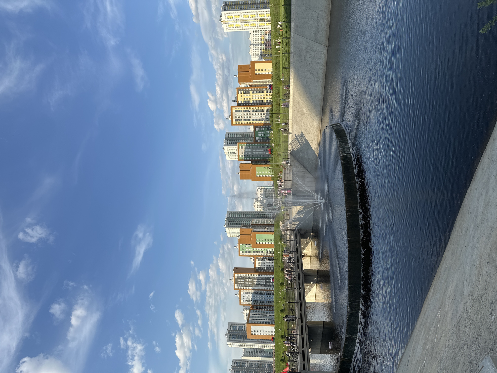 This image shows a portion of the Yekaterinburg City Pond (Gorodskoy Prud) in Yekaterinburg, Russia, with a concrete embankment in the foreground, and a view across the pond towards high-rise apartment buildings. The sky is blue with scattered white clouds. A multi-tiered artificial waterfall cascades into the dark water of the pond. Pedestrians are visible walking along the embankment.